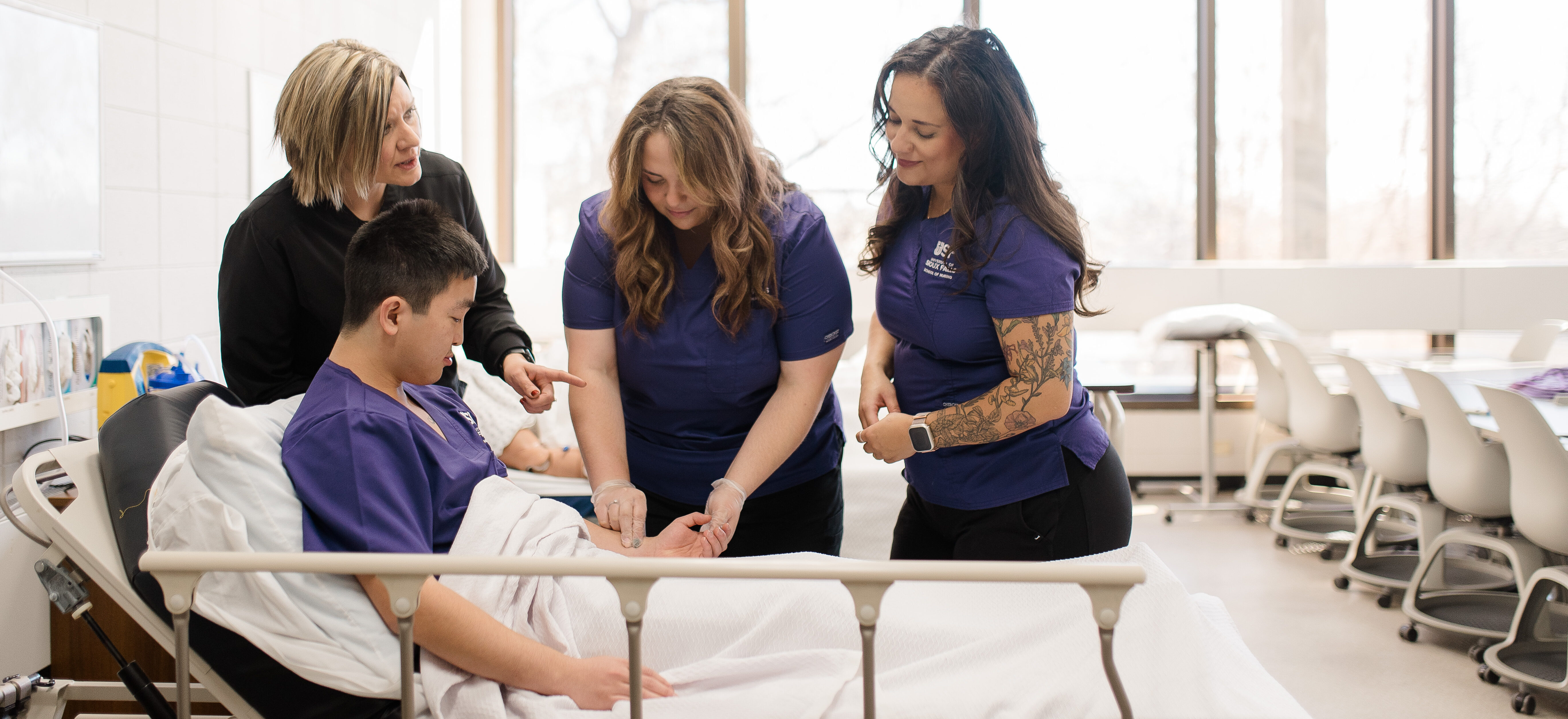 Four people surround a bed-ridden patient in a brightly lit room. Three women in purple uniforms attentively focus on the patient, one holding a medical device. Another woman, in black, appears to provide instruction, creating an atmosphere of care and learning.