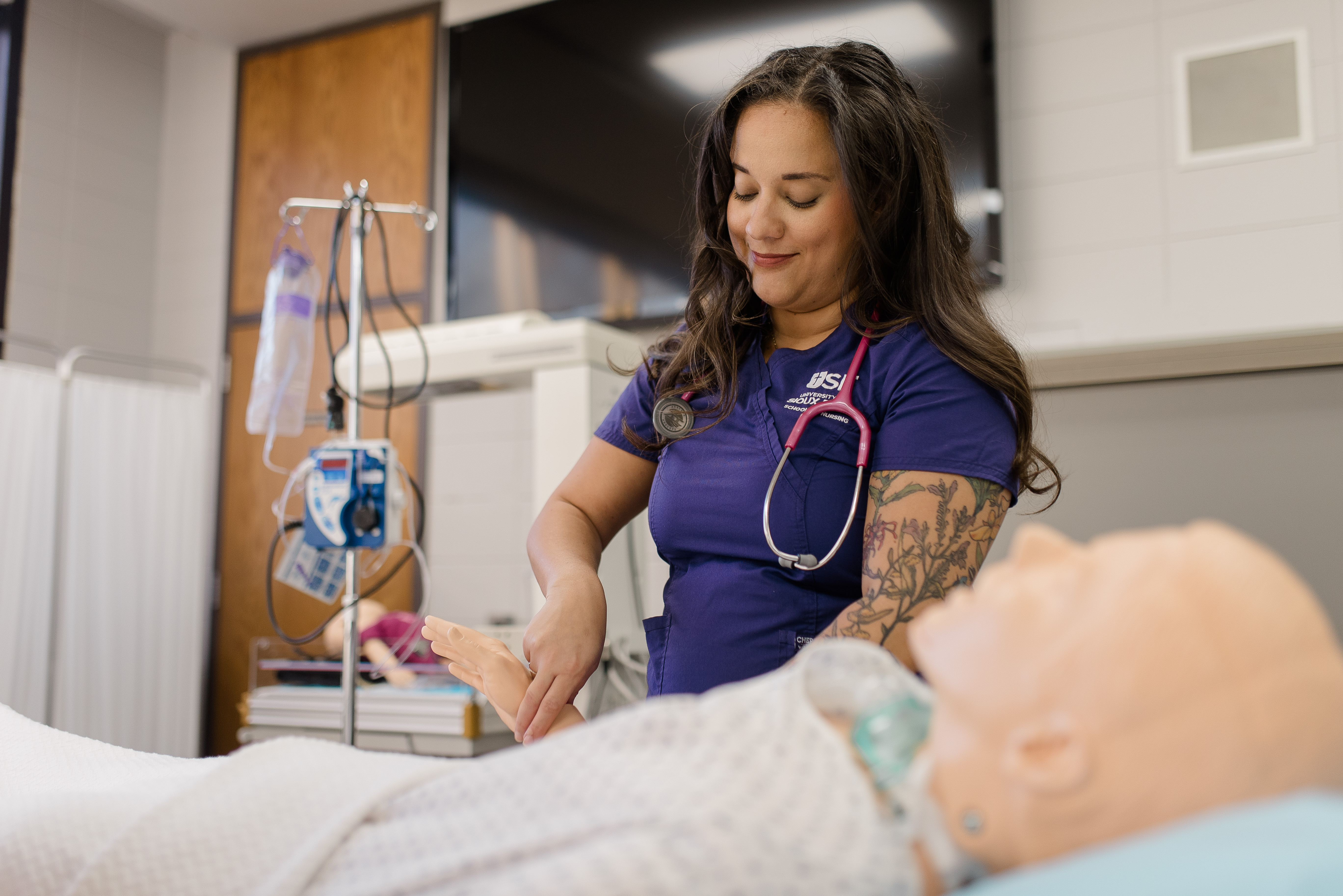 A healthcare professional in a blue uniform and stethoscope around her neck is attending to a medical mannequin in a hospital training room. She is smiling and holding the mannequin's hand. Medical equipment and a privacy screen are visible in the background.