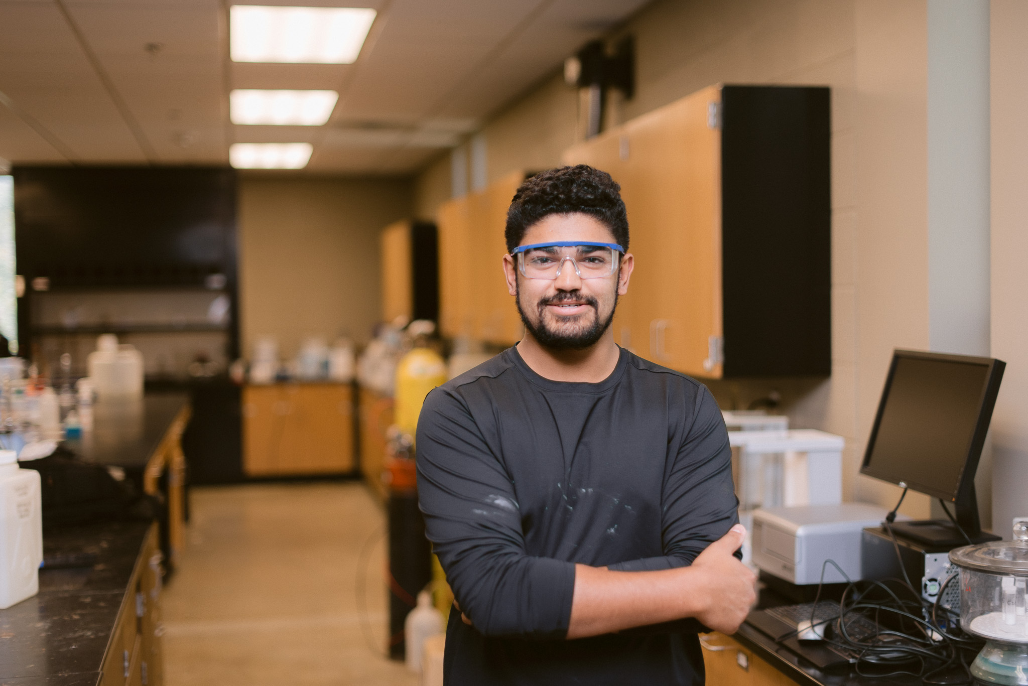 A man wearing safety glasses and a black long-sleeve shirt stands confidently with arms crossed in a laboratory. The lab has wooden cabinets, countertops with various equipment, and a computer setup on the right. The atmosphere appears bright and professional.