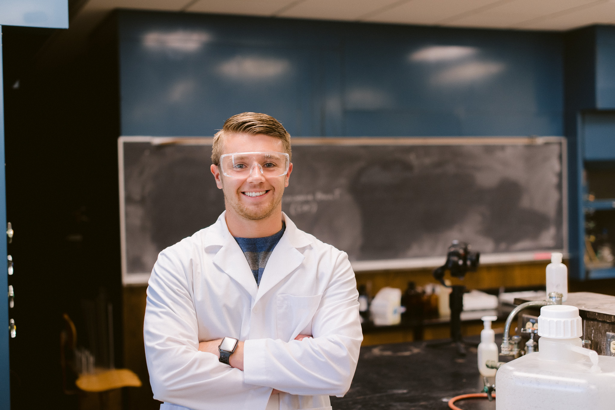 A person wearing a white lab coat and safety goggles stands with their arms crossed in a science laboratory. The background features a chalkboard and various scientific equipment. The person is smiling and appears to be confident and engaged in their work.