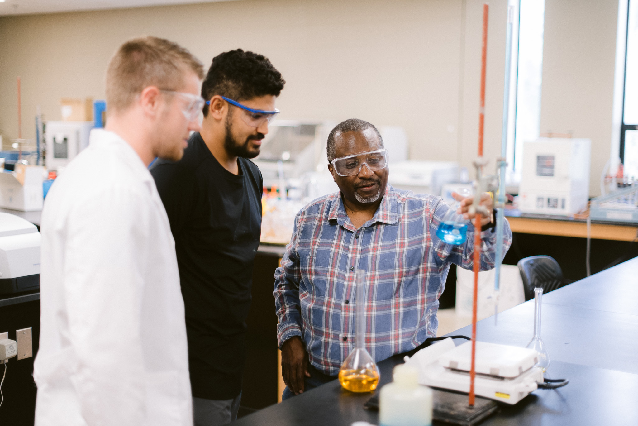 Three people in a laboratory, wearing safety goggles. The person on the right, in a plaid shirt, holds a beaker with blue liquid while explaining something. The other two people, one in a white lab coat, look on attentively. Lab equipment is visible in the background.