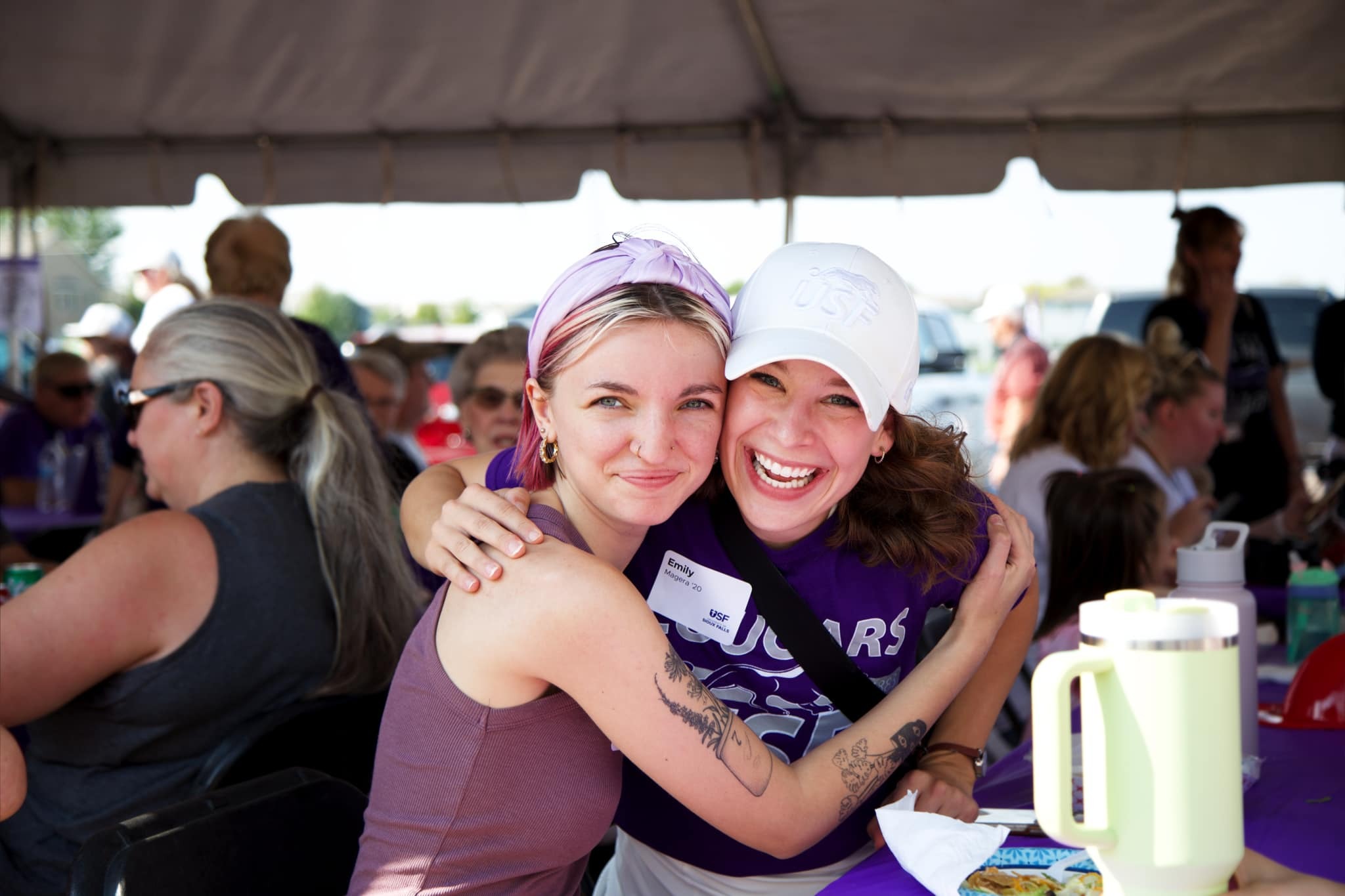 Two women at an outdoor event smile and hug under a tent. One wears a light pink headband with a tank top and the other sports a white cap and a purple shirt. People are seated at tables in the background, indicating a social gathering.