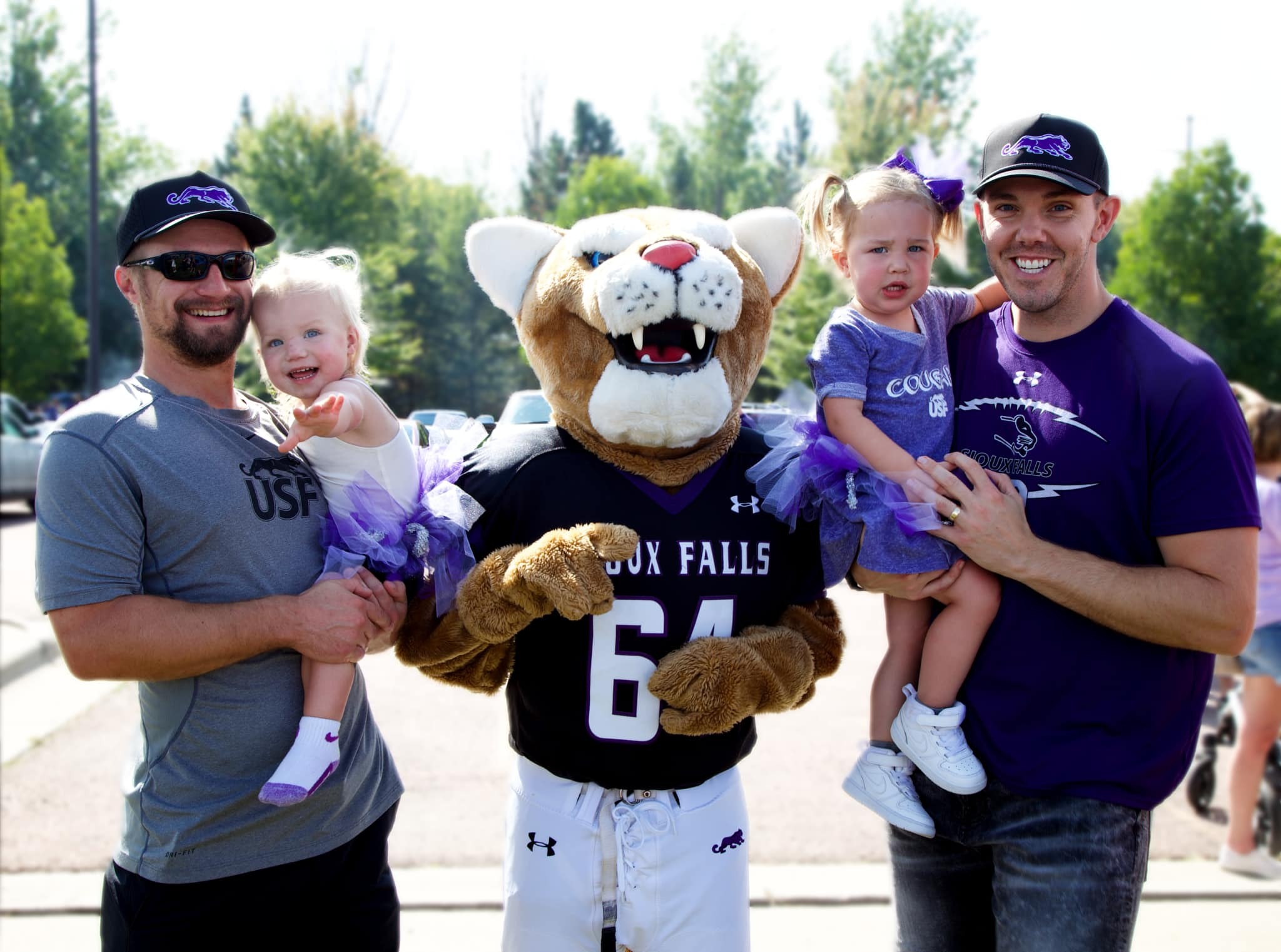 Two men, each holding a young child, stand on either side of a sports mascot dressed in a cougar costume with the number 64 and "Cougars" on the jersey. The men and children are smiling, and the children are dressed in matching outfits with purple tulle skirts.