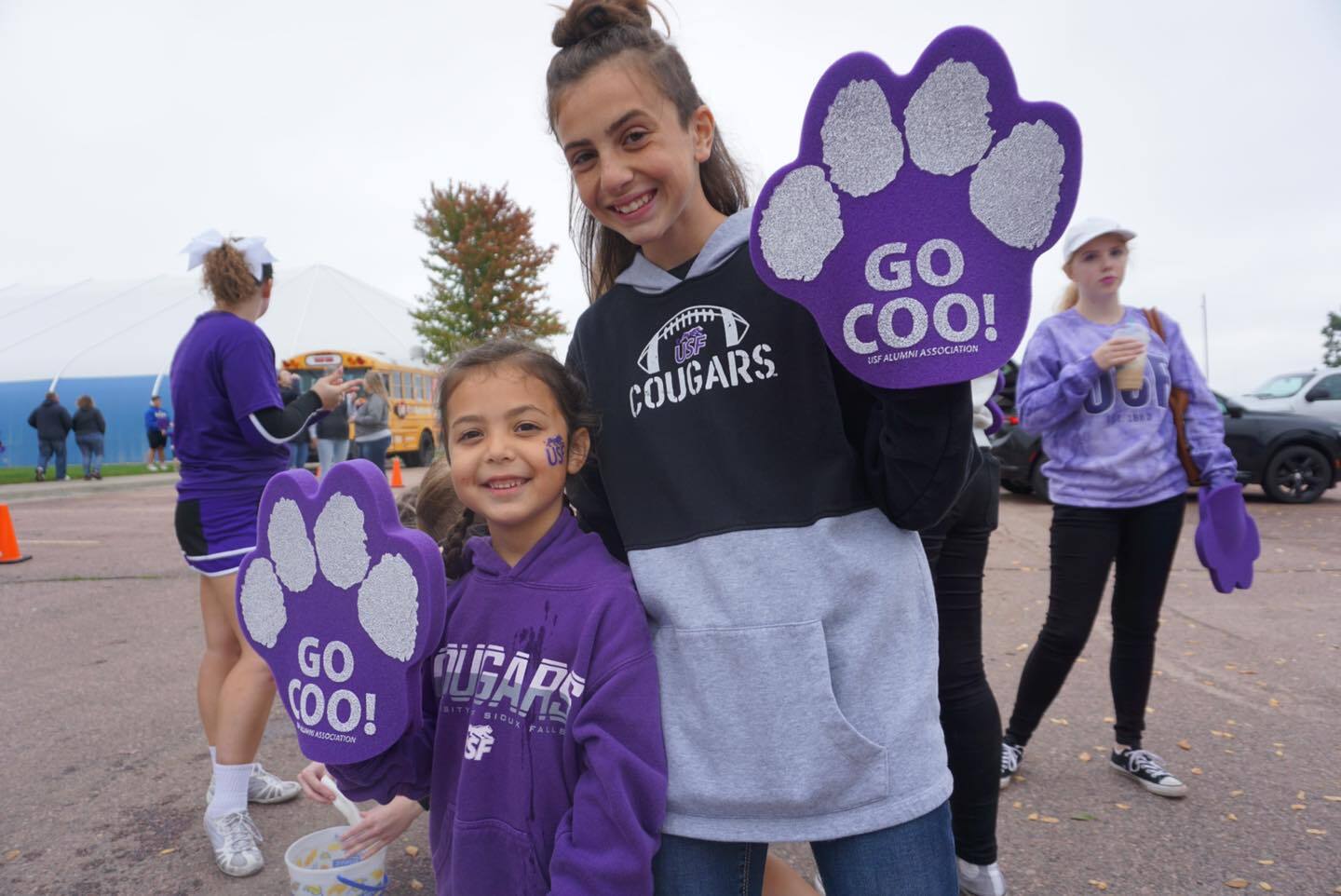 Two smiling girls pose during a school spirit event. Both hold up large foam paw prints that read "GO COO!" while dressed in Cougar-themed clothing. The background shows other participants and a blue building. One girl eats a snack, and the ground is dotted with leaves.