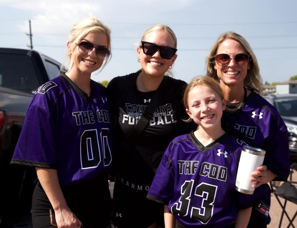 Four smiling people are posing together outdoors. Three are wearing matching purple football jerseys with "The COD" and different numbers, while the fourth person, dressed in black, has "Cougars Football" on their shirt. One holds a metal tumbler. They appear to be enjoying a sunny day.