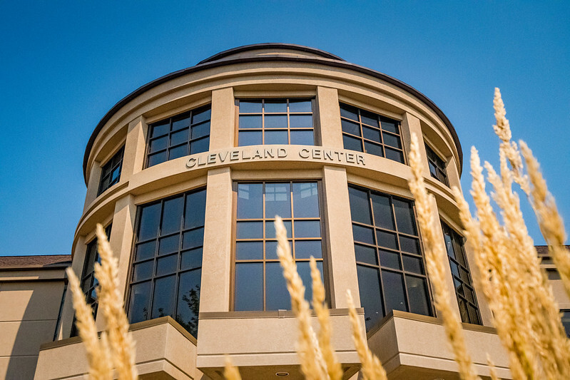 A modern building with large windows and a rounded facade labeled "Cleveland Center." The building is set against a clear blue sky, with tall golden ornamental grasses in the foreground.