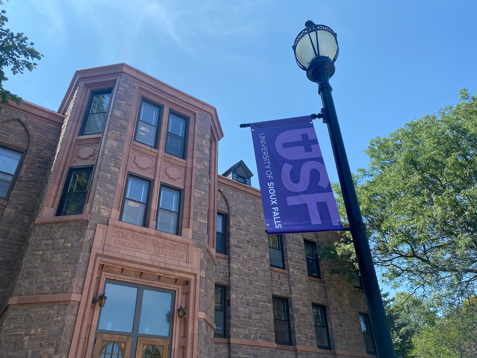 A building made of stone and bricks with arched wooden doors, shown under a clear blue sky. A banner on a lamppost reads "USF University of Sioux Falls" in purple with white text. Trees and greenery are visible around the area.