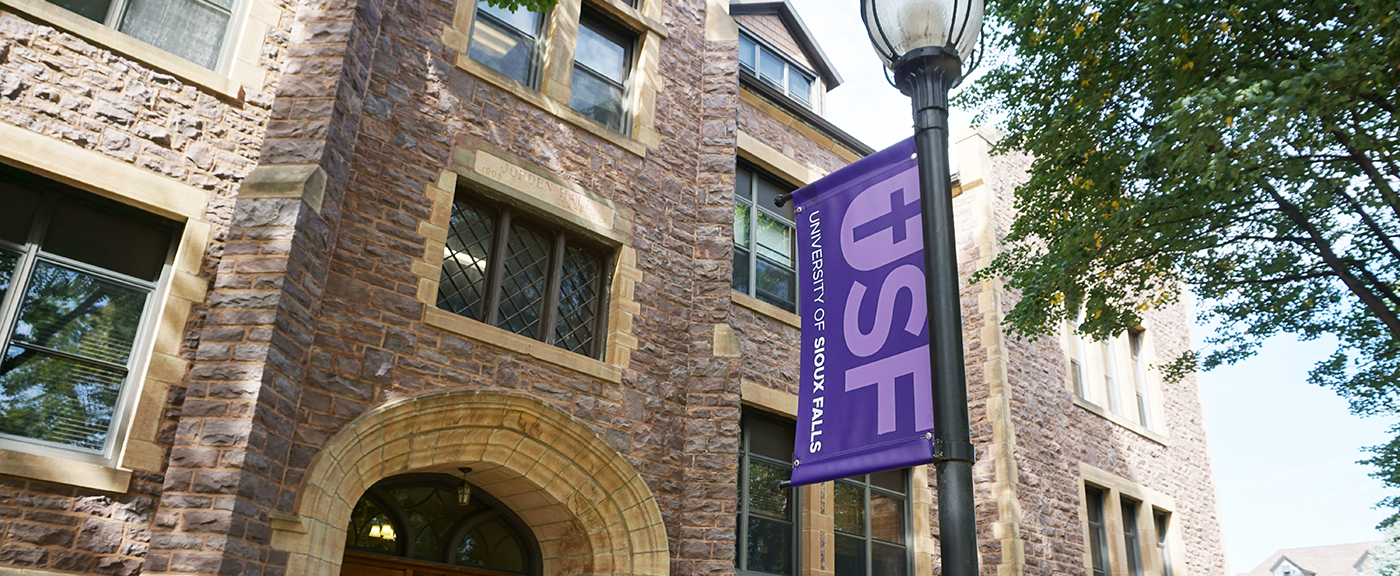 An old, multi-story building constructed with rustic stone features arched windows and a wooden double-door entrance. A purple banner displaying "USF University of Sioux Falls" hangs from a nearby lamppost. Trees with green leaves surround the building.