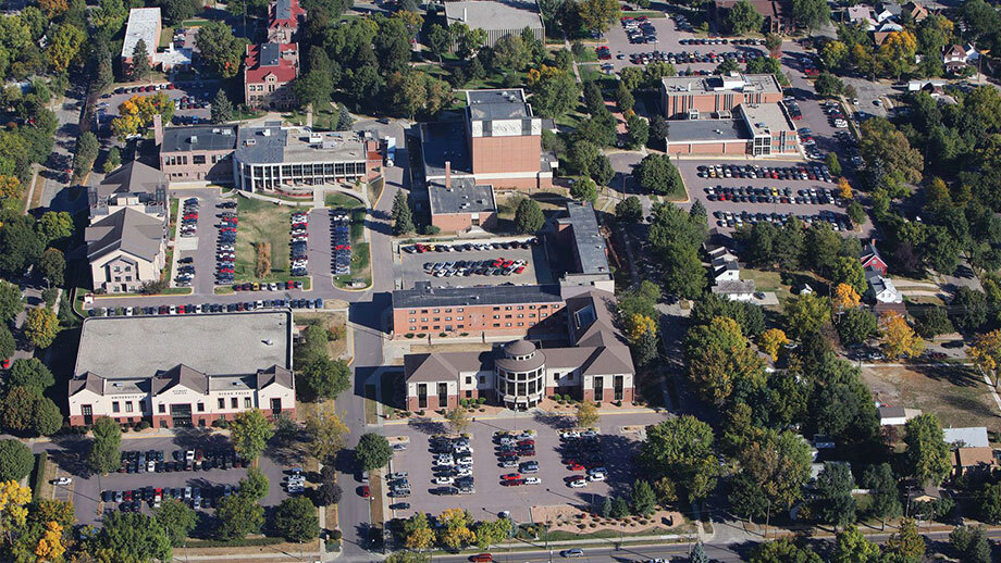 Aerial view of a college campus with multiple buildings surrounded by parking lots and trees. People and cars are visible throughout the scene. The architecture includes a mix of modern and older styles, with a central circular building standing out.