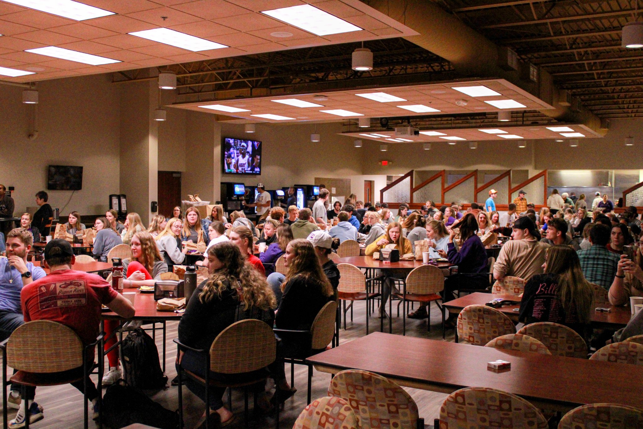 A bustling cafeteria filled with people sitting at tables, eating, and talking. Some are standing in line at food stations. The room has a high ceiling with exposed beams and fluorescent lighting. A TV is mounted on the wall in the background showing a sports game.