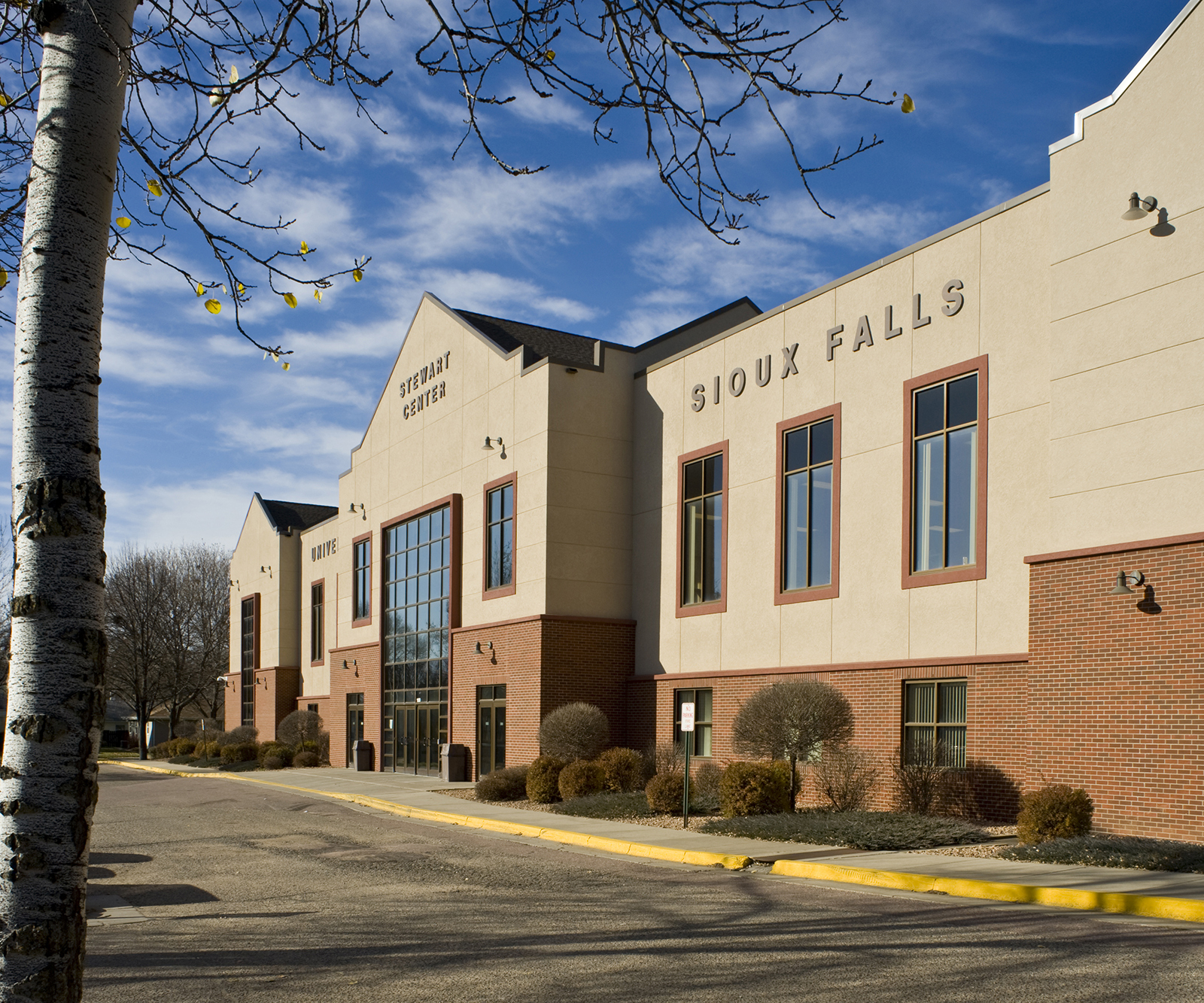 A building with a prominent entrance and large windows, labeled "Sioux Falls" and "Event Center." The structure has beige and brick exteriors, with a few small trees and shrubs lining the walkway. A leafless tree is in the foreground, with a clear blue sky overhead.
