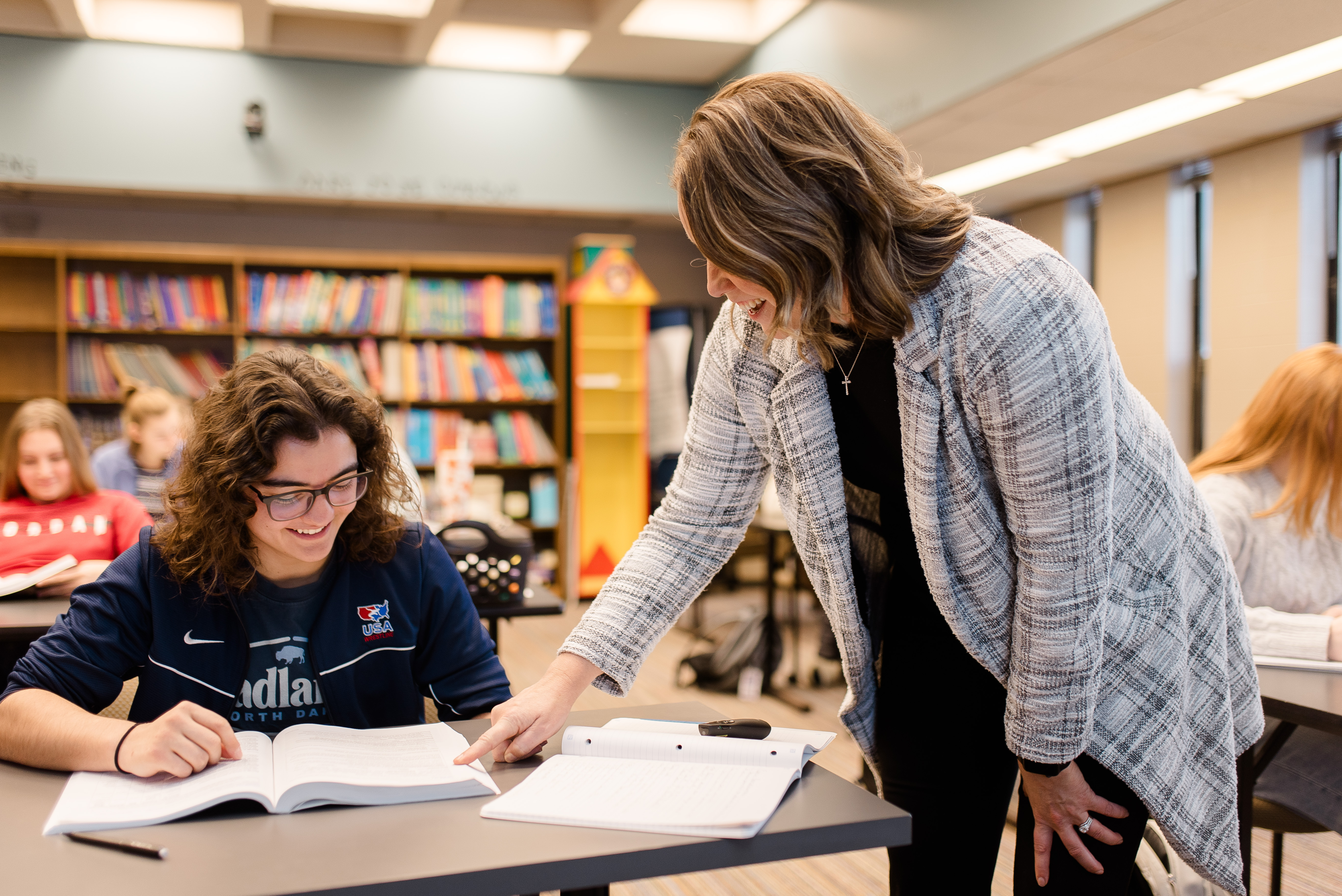A teacher stands next to a seated student in a classroom, pointing to a page in the student's open book. The student is smiling, and other students are visible in the background. The classroom has bookshelves filled with books.