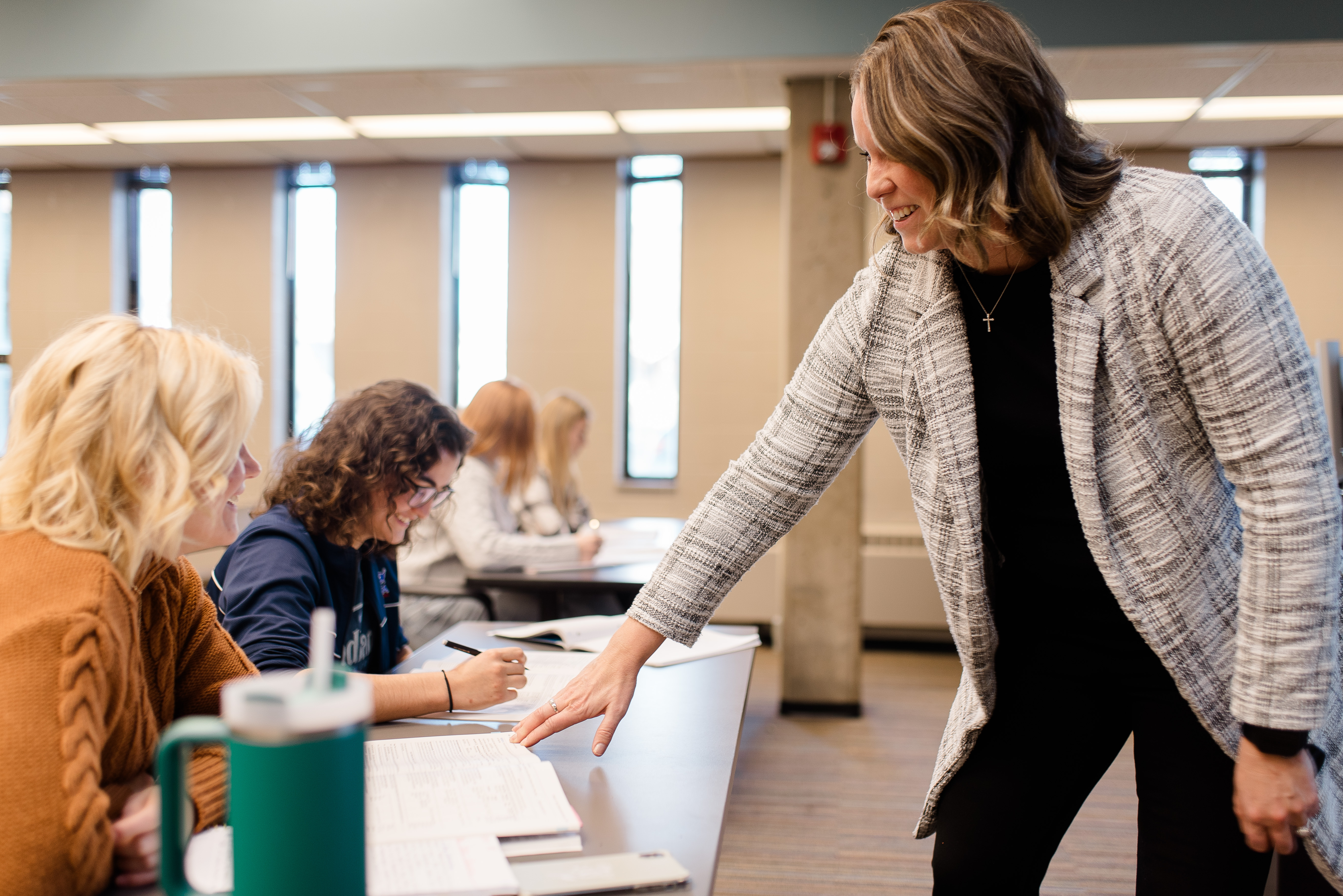 A woman stands by a desk in a classroom, smiling and pointing to a document that a student is working on. Other students are seated at the desk, focusing on their work. The classroom has large windows letting in natural light.
