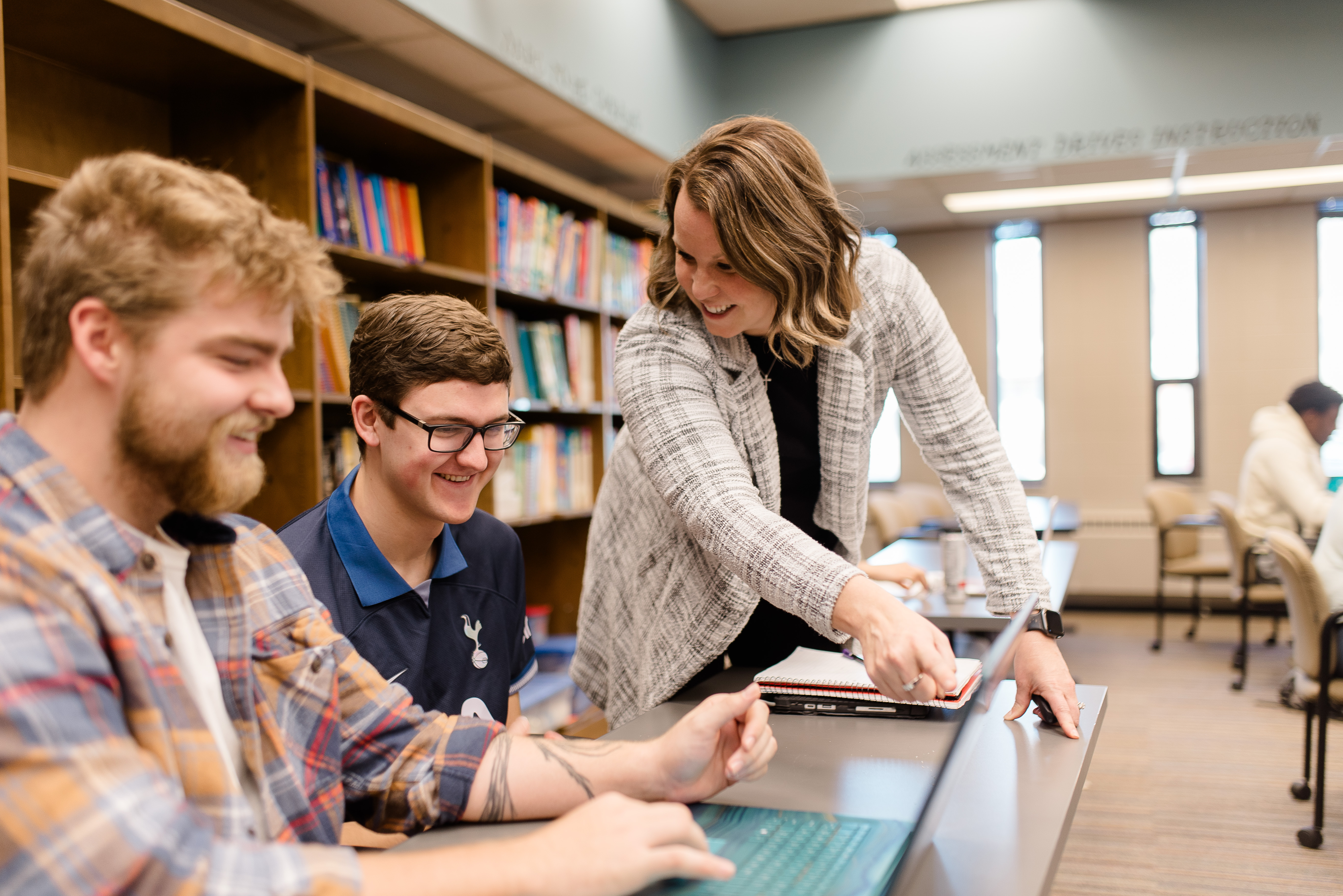 A woman stands beside two smiling men seated at a table in a library. The woman points to a spot on a paper while the men look on. Shelves filled with books line the background. Other students are visible studying at tables further back.