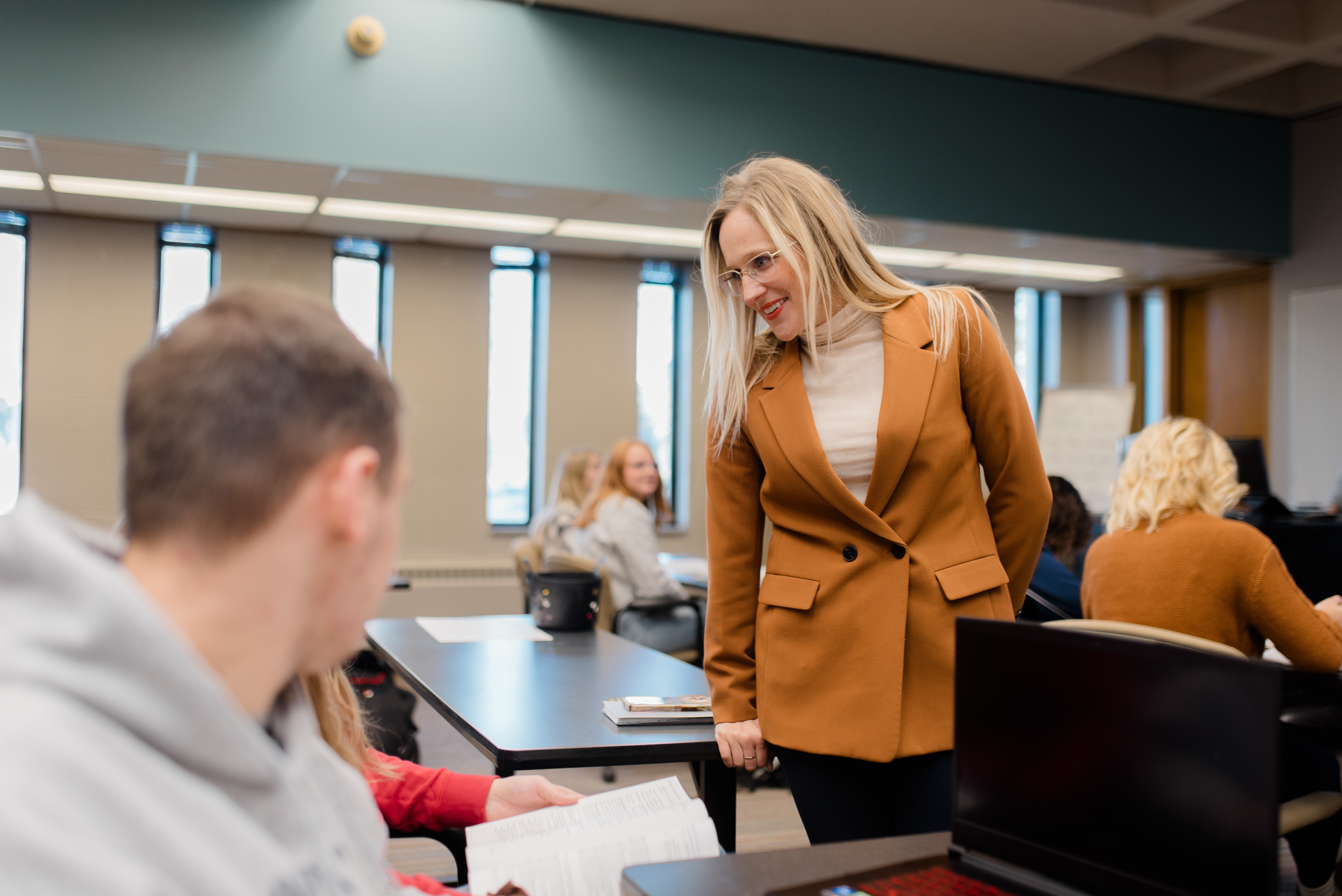 A woman in a brown blazer and glasses is standing and engaging with a seated person in a classroom. Other students are sitting at tables in the background, with books and laptops on the desks. The room has large windows with blinds partially open.