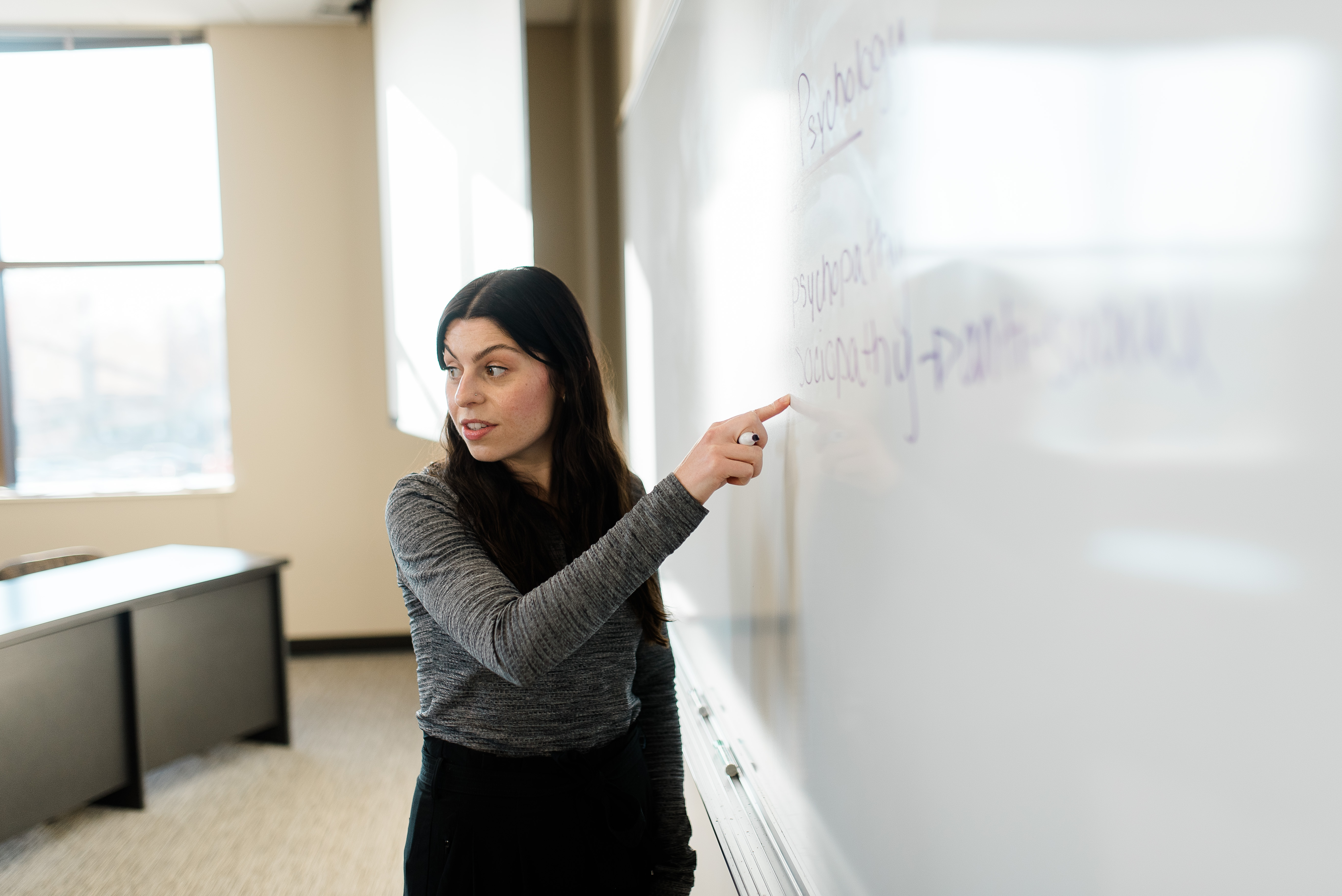 A person with long dark hair, wearing a gray long-sleeve shirt, is pointing to something written on a whiteboard in a classroom. The classroom is bright with natural light coming through the windows. The individual appears to be explaining or teaching.