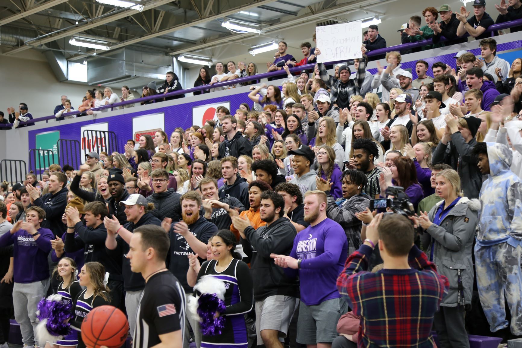 A large group of enthusiastic supporters, many dressed in purple and holding up signs, cheers from the bleachers at a sporting event. Cheerleaders in uniform hold pompoms in front, while a player with a basketball is seen in the foreground.