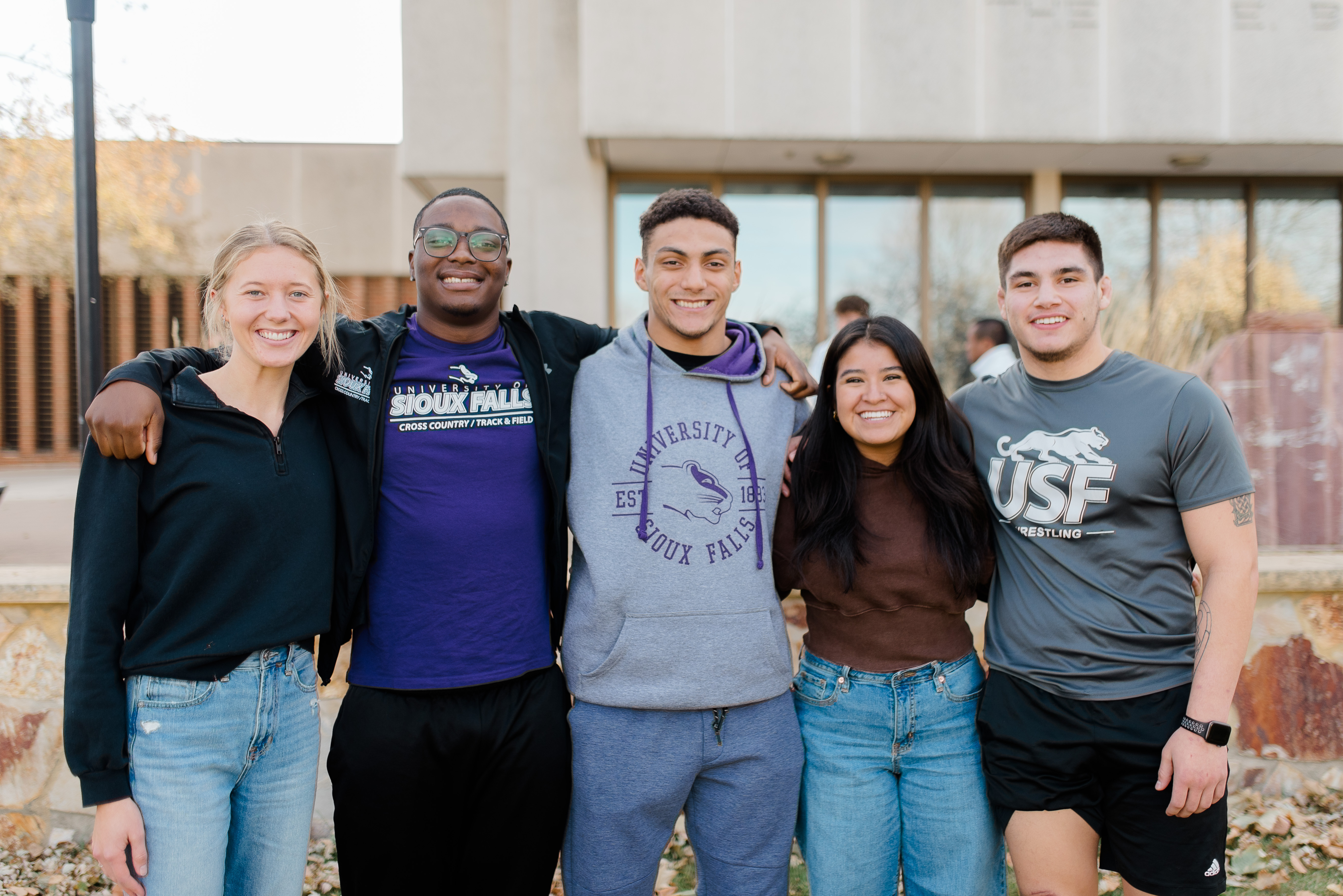 A group of five people stand close together outdoors, smiling at the camera. They appear to be college students and are casually dressed, with some wearing University of Sioux Falls apparel. A building and trees are visible in the background.