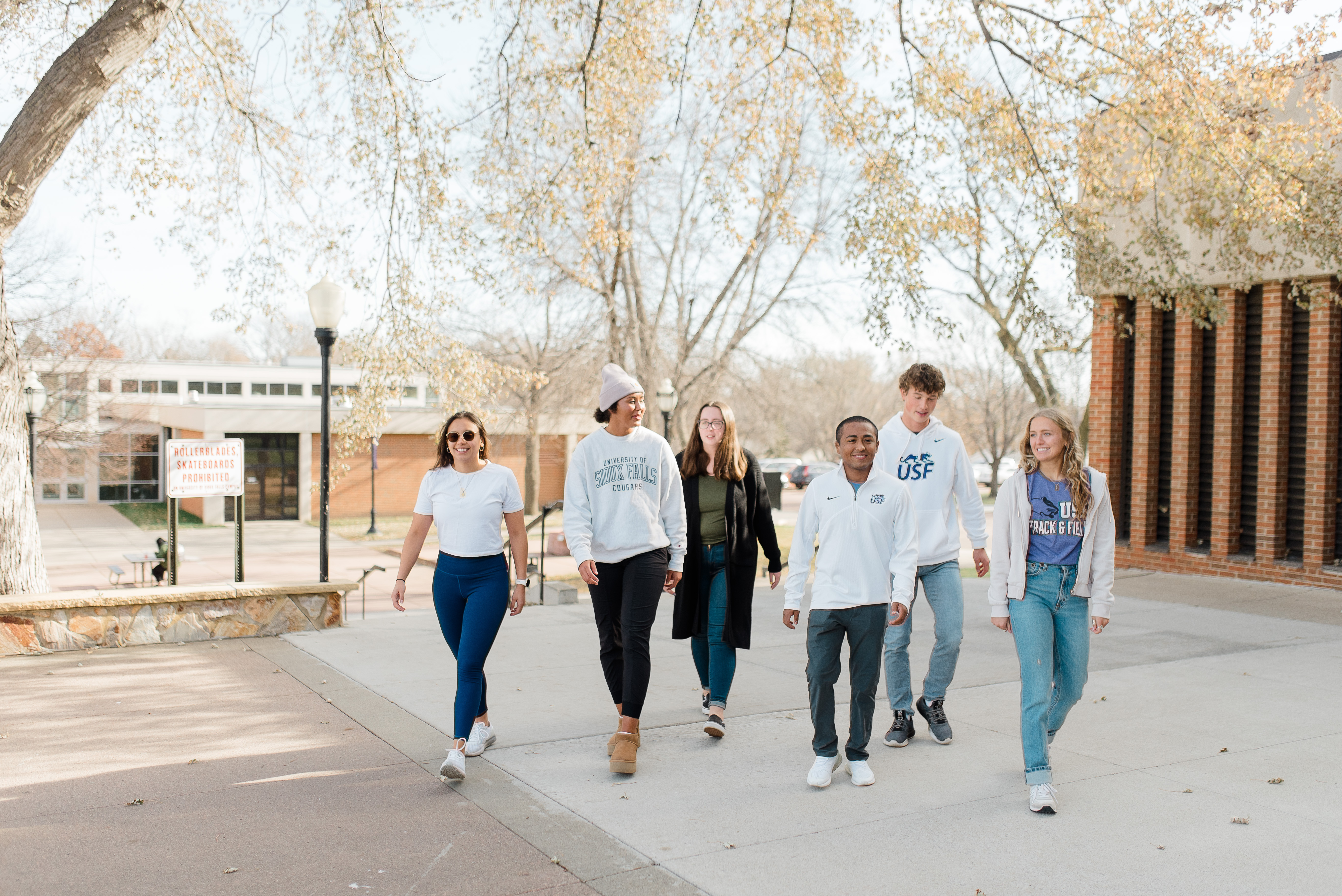 A group of six young adults, dressed casually, walks outside on a sunny day. They laugh and chat amongst themselves. Trees with sparse autumn leaves and campus buildings are visible in the background.