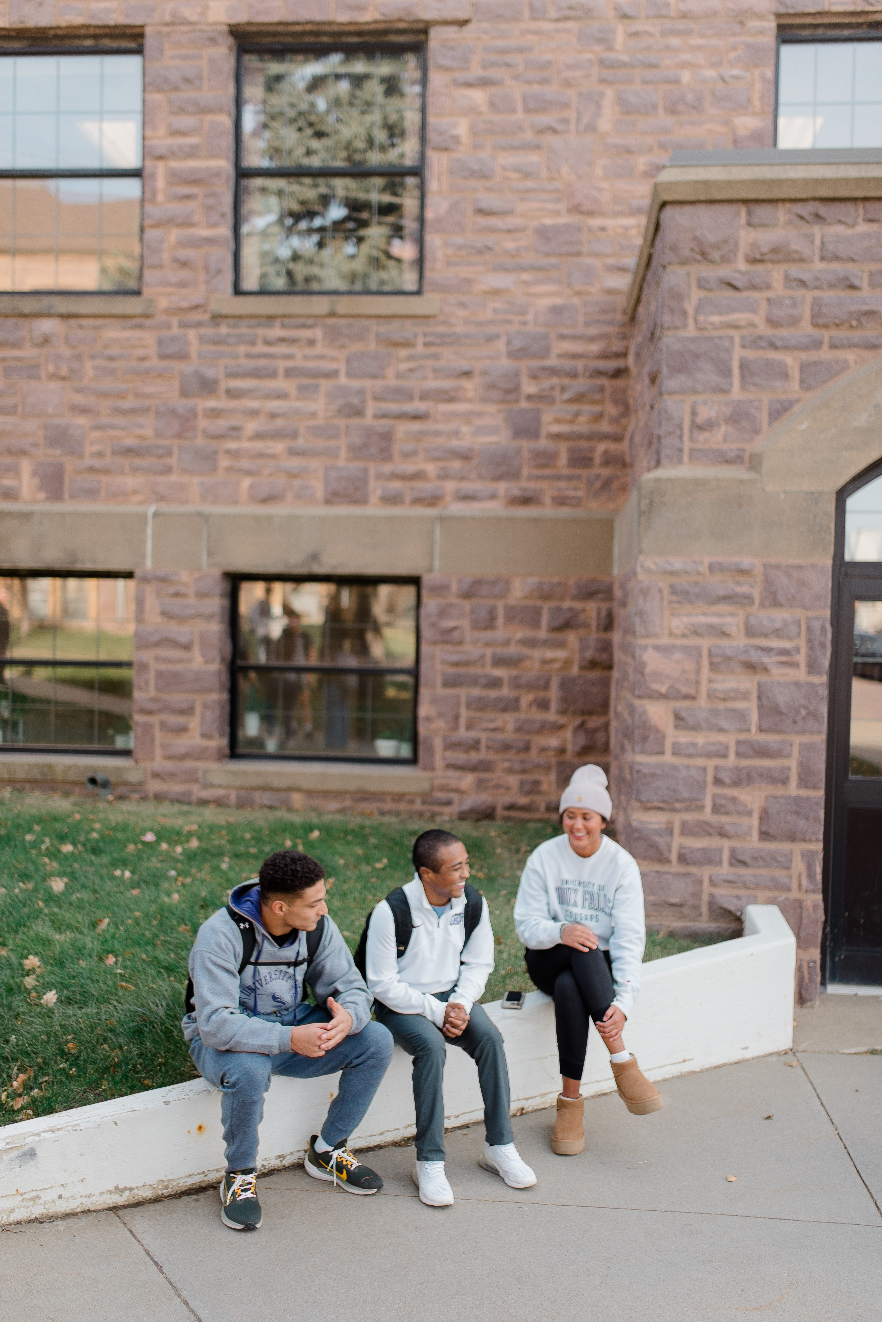 Three friends sit on a white ledge outside a stone building with large windows. Two of them are looking at the third person, who is smiling and wearing a white beanie. There's green grass behind them, and the scene appears to be casual and relaxed.