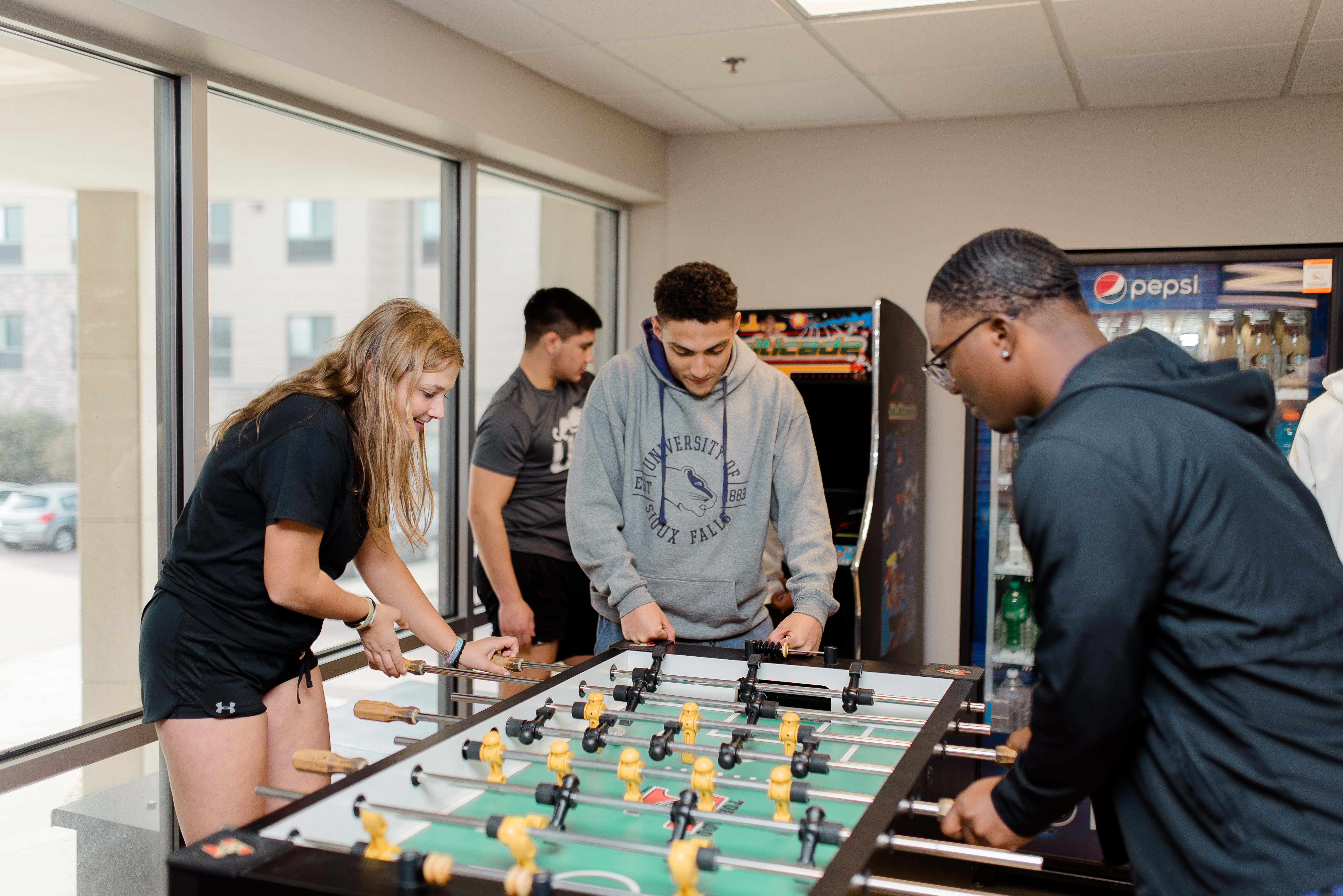 Four people are casually playing foosball in a well-lit indoor space with large windows. The group appears engaged in the game, with soft drinks visible in the background. Behind them, a Pepsi vending machine and a soft drinks display can be seen.