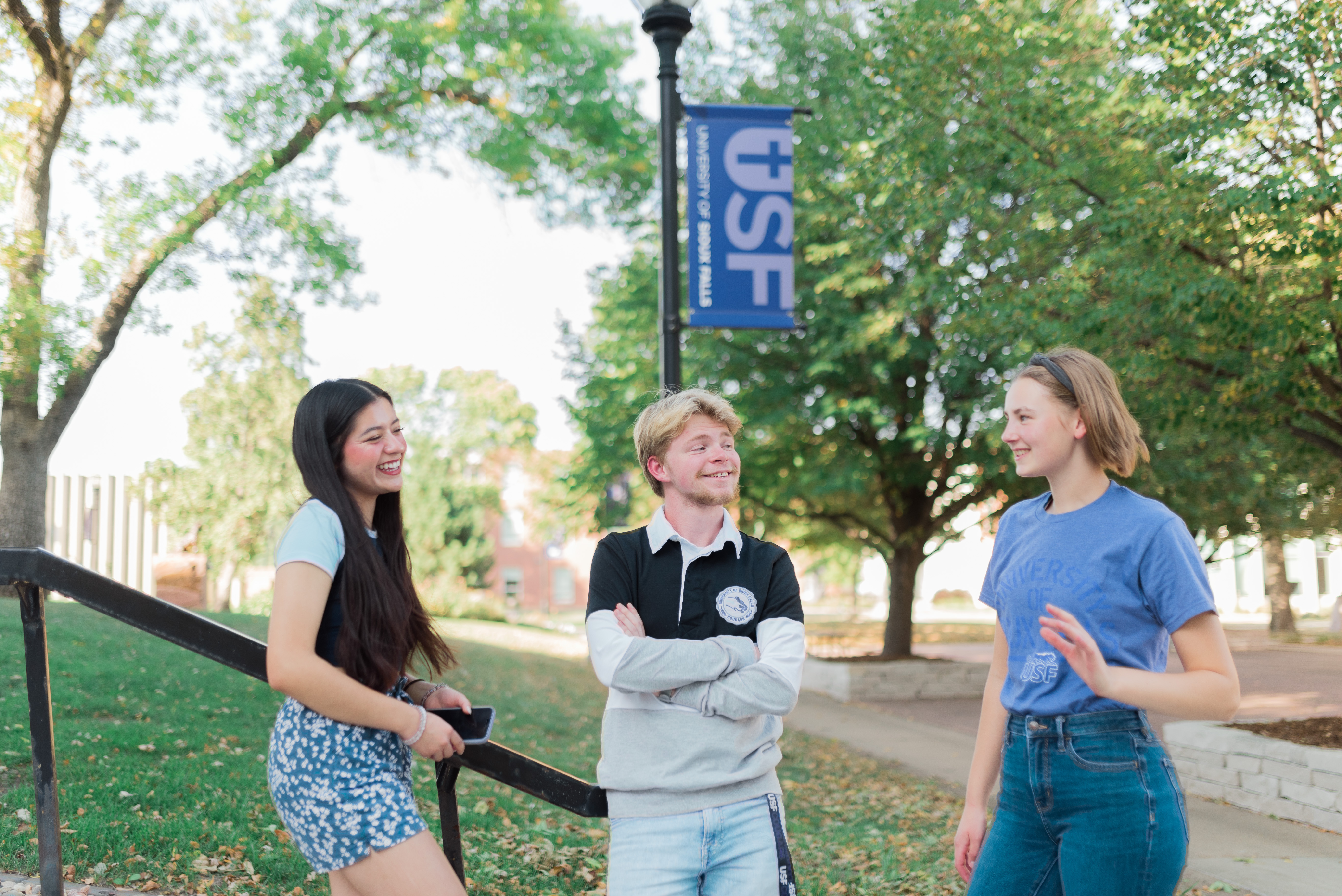 Three college students are standing outdoors on a sunny day, laughing and talking. Behind them, there are leafy green trees, a paved walkway, and a tall lamppost with a banner that reads "USF." The students are casually dressed and appear to be enjoying their conversation.
