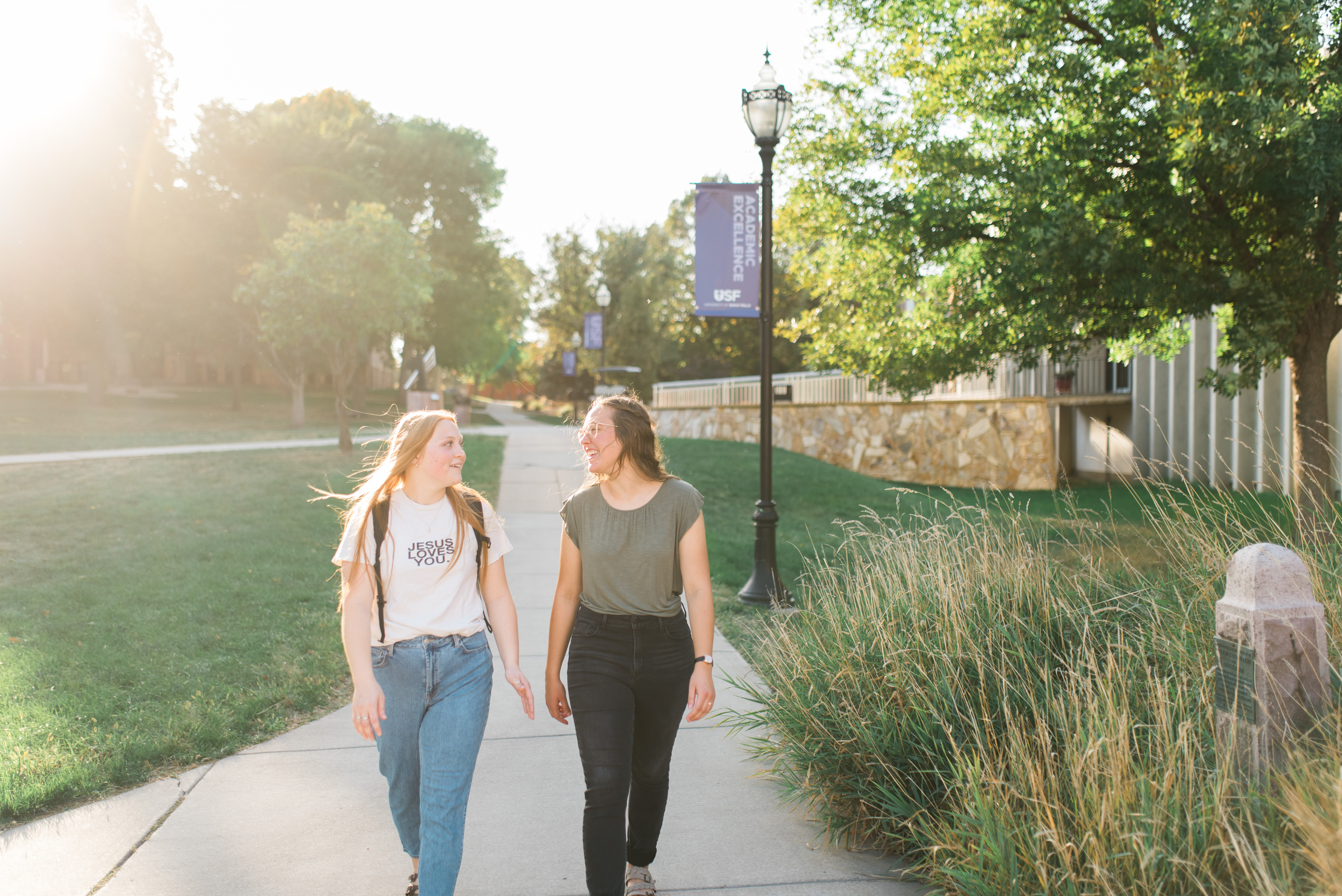 Two people walk down a sunlit path on a college campus, smiling at each other. One wears a white T-shirt and jeans; the other wears a green top and black pants. They are surrounded by green trees and grass, with a light post and banner visible in the background.