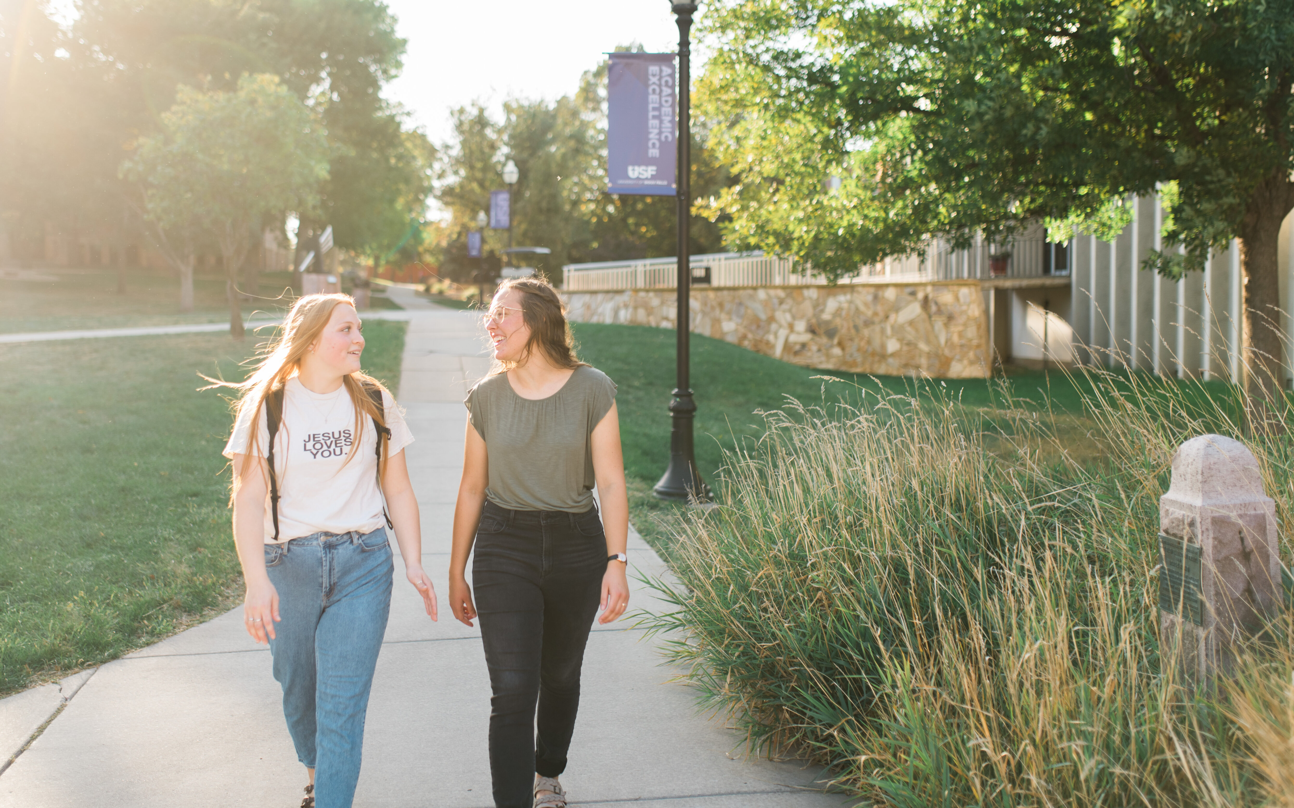 Two young women walk along a sunlit path on a college campus. One wears jeans and a white t-shirt with "JESUS LOVES YOU" printed on it, while the other wears dark pants and a green top. They smile at each other, surrounded by greenery and a lamppost displaying a banner.