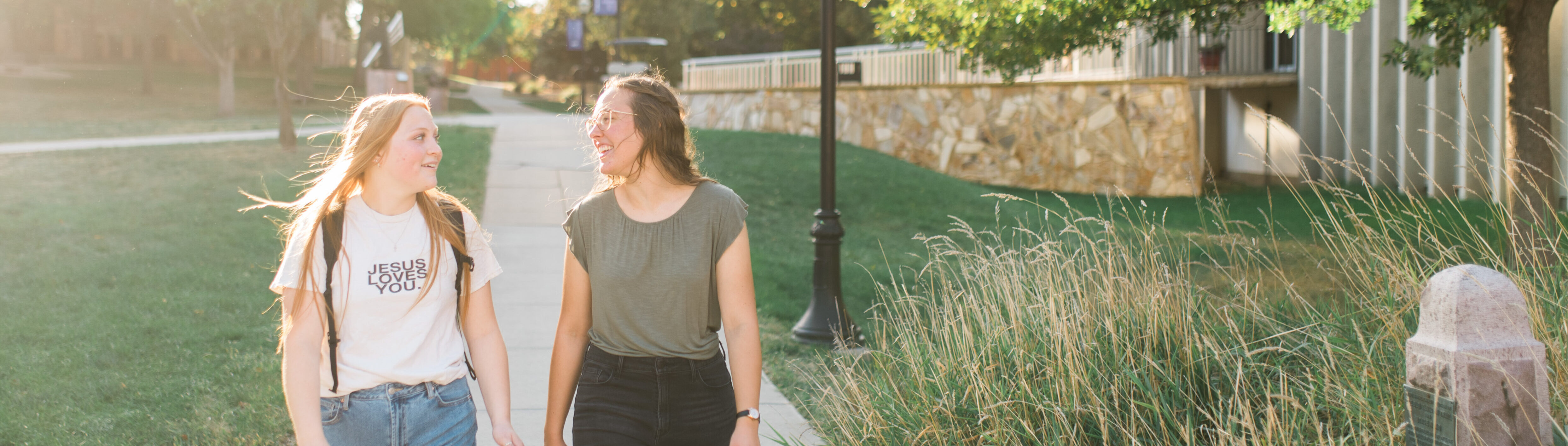 Two young women walk along a sunlit sidewalk, smiling at each other. One wears a white T-shirt with "JESUS LOVES YOU!" printed on it and carries a backpack, while the other wears a green T-shirt. They stroll beside greenery and a building.