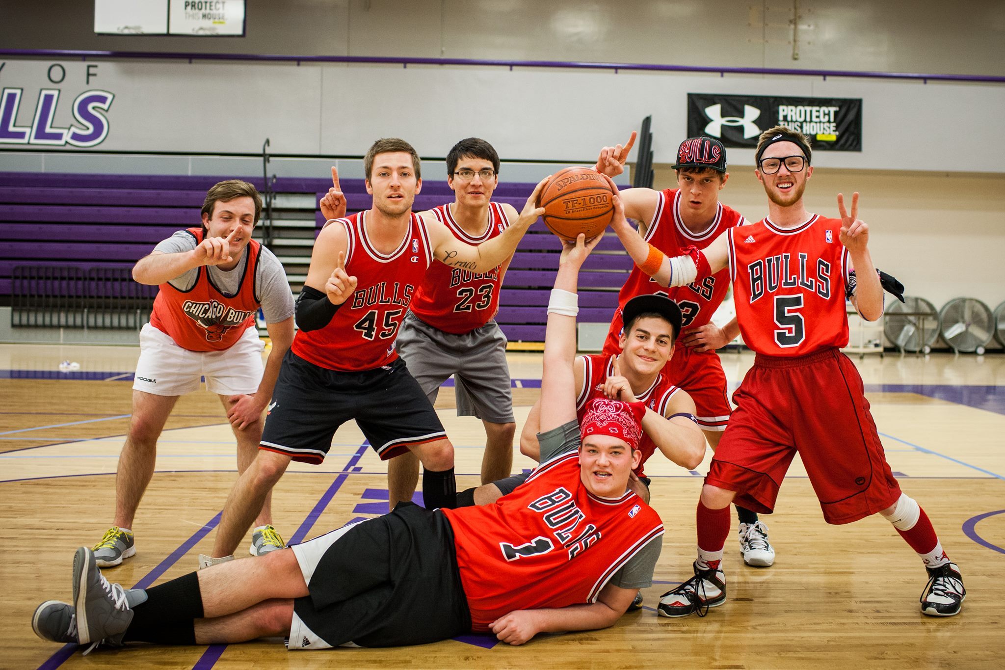 A group of seven young men poses on a basketball court, all wearing assorted Chicago Bulls jerseys. One person is holding a basketball above others while another lies on the floor. Some are making peace signs with their fingers. The court has purple accents.