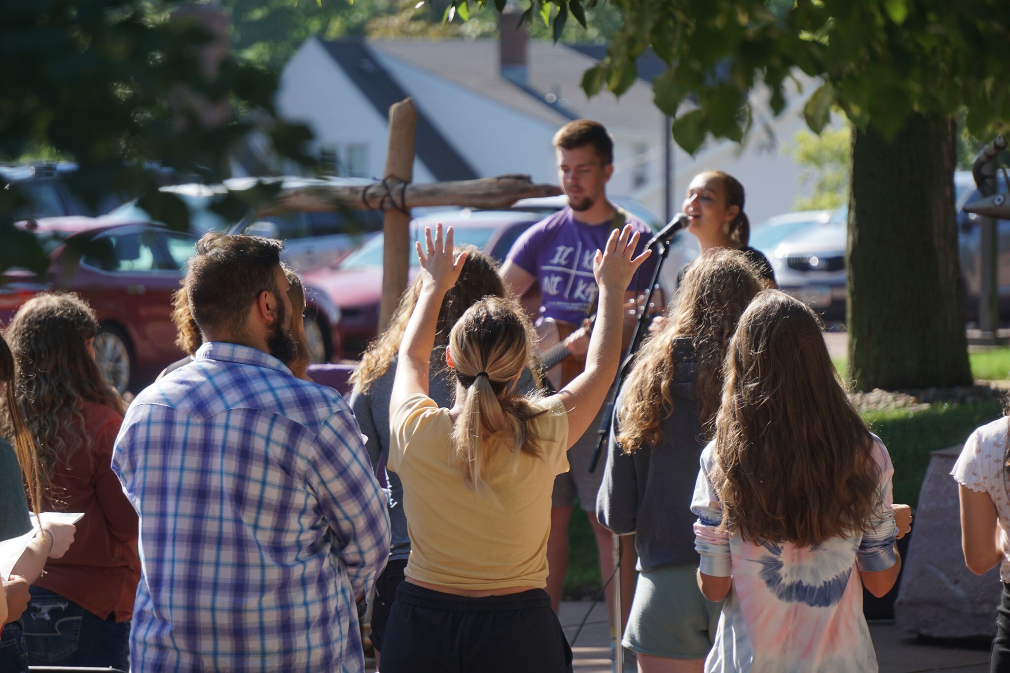 A group of people facing a stage with a man and a woman singing into microphones. One person in the foreground has their hands raised. The gathering is outdoors with trees and cars visible in the background.