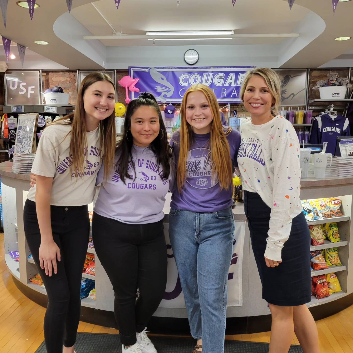 A group of four smiling people stand inside a store with "COUGARS" banners and merchandise. Three are wearing University of Sioux Falls (USF) themed shirts, and one person, standing to the right, is dressed in a white blouse and dark skirt.