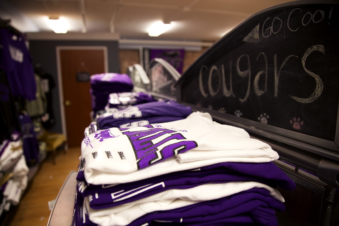 A small retail store with neatly folded stacks of purple and white sports apparel on shelves. In the background, a chalkboard sign reads "GO COUGARS" with small paw prints drawn next to the text. The store has a cozy and organized ambiance.