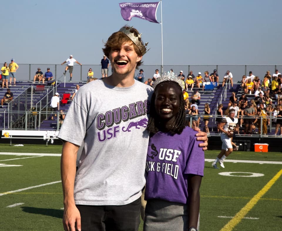 Two students stand on a football field, smiling and wearing crowns. They are wearing grey and purple shirts with "USF" written on them. Fans in the background are sitting on bleachers, and there is a player in a football uniform behind them.