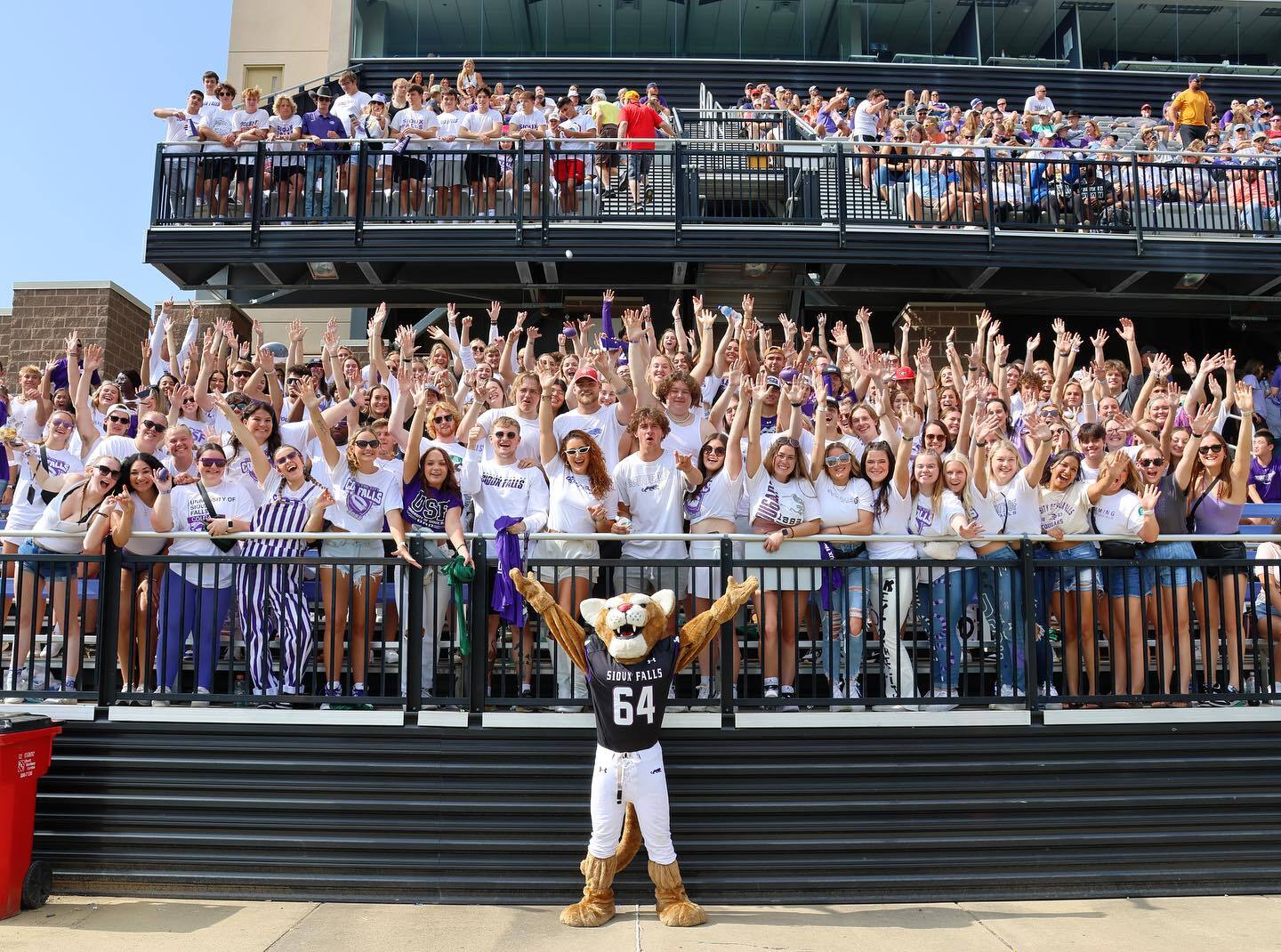 A costumed mascot wearing a jersey with the number 64 is standing in front of a large crowd of cheering people. The crowd, dressed mostly in white and purple, are gathered on a platform and waving their hands in the air. Everyone appears to be celebrating outdoors.