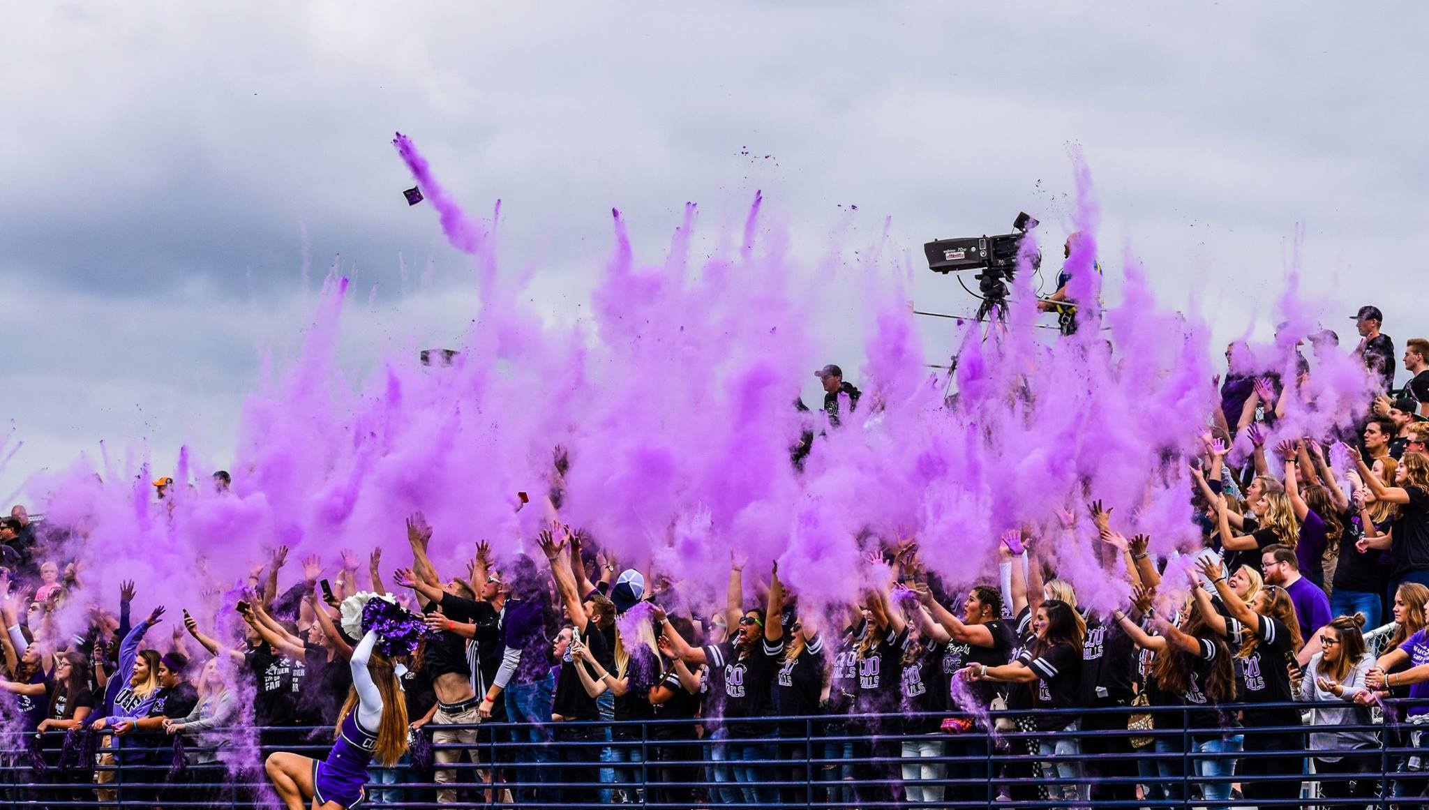 A large crowd of enthusiastic fans dressed in matching colors throw clouds of purple powder into the air, creating a vibrant and energetic atmosphere. The sky is overcast, and some spectators hold their hands up while others cheer.