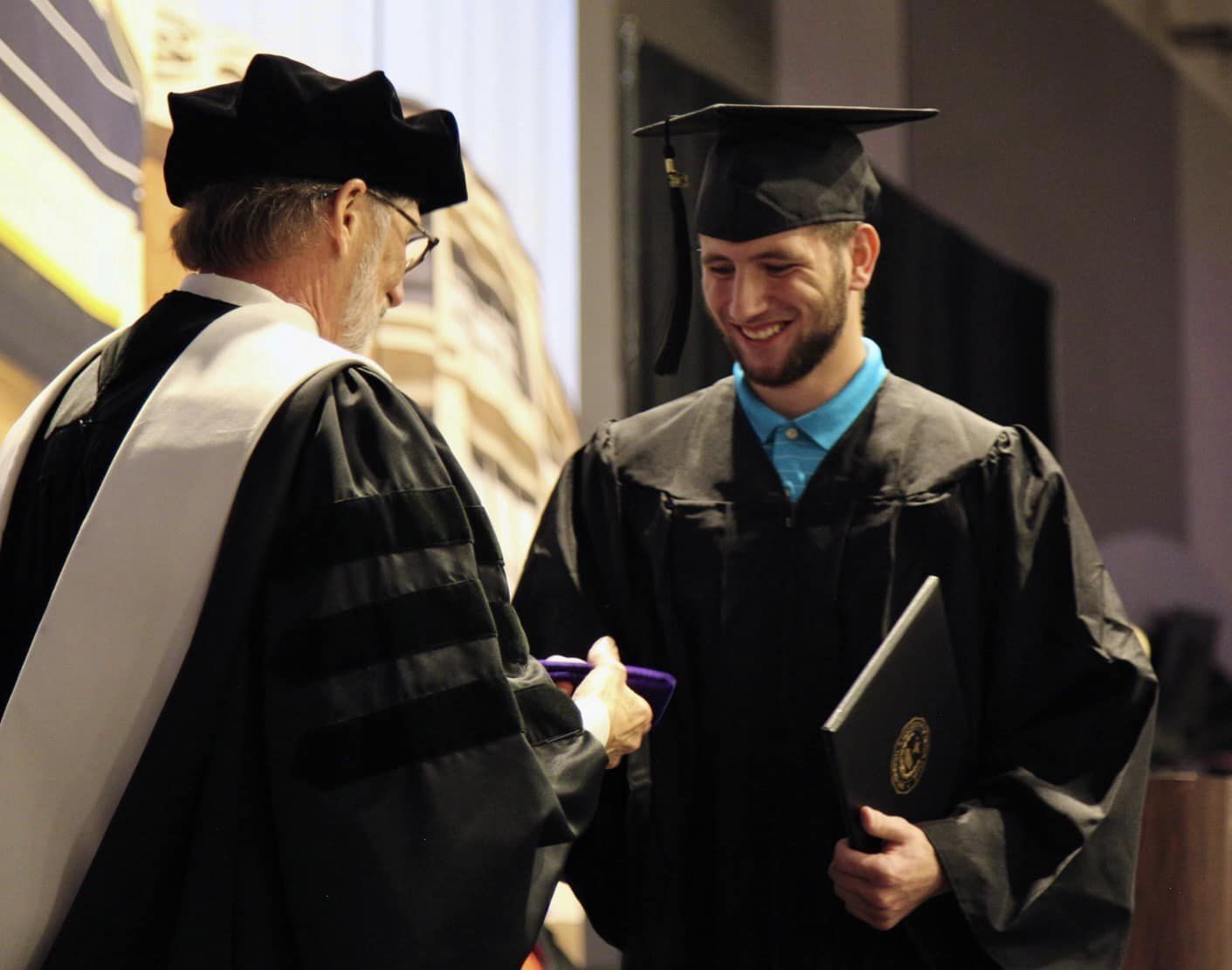 A graduate in a cap and gown smiles as he receives his diploma from a faculty member wearing academic regalia during a graduation ceremony. The faculty member is handing over the diploma with both hands while the graduate holds it with his left hand.