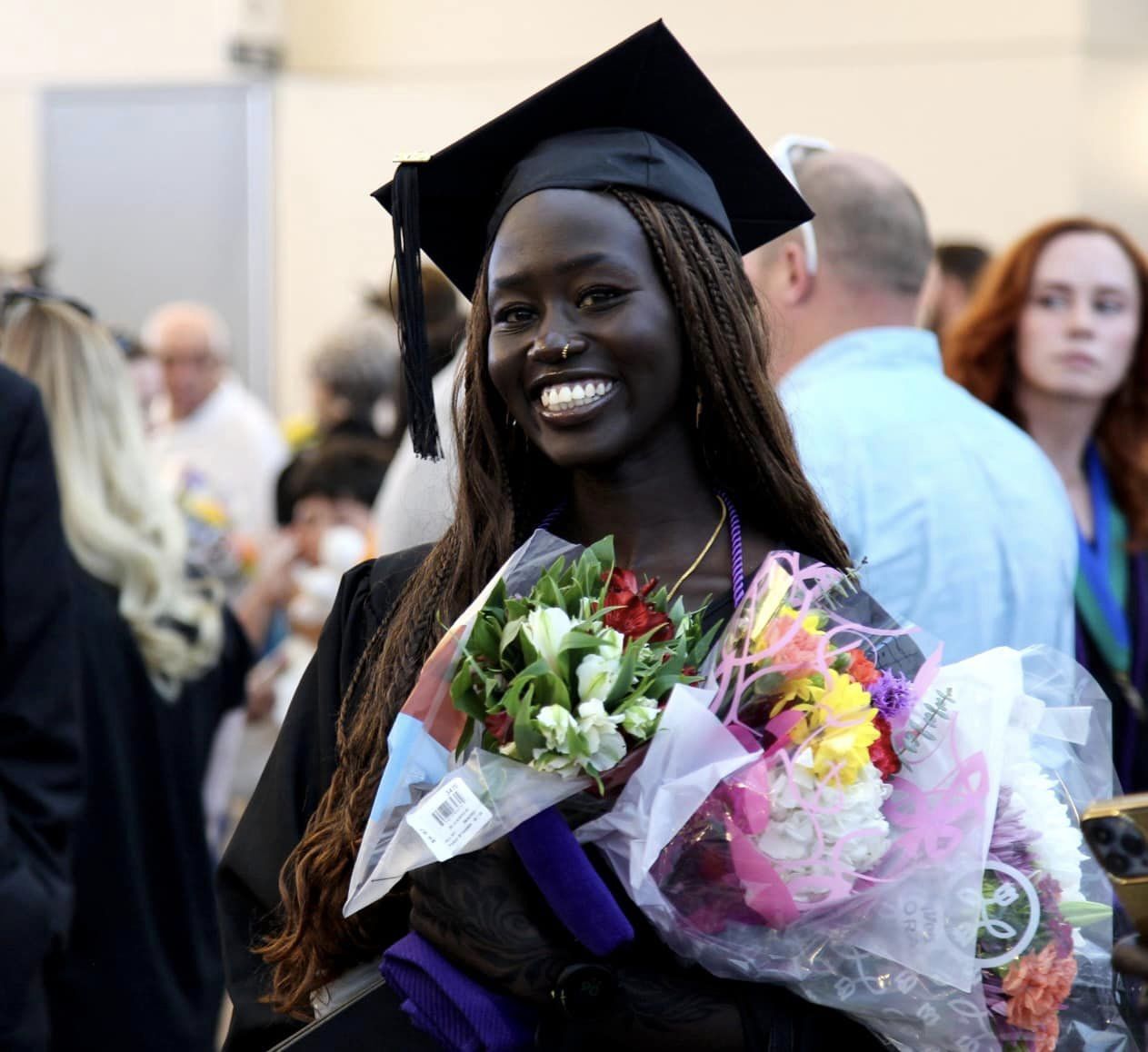 A graduate in a black cap and gown holds a bouquet of flowers and smiles broadly at the camera. Other people and graduates are visible in the blurred background, suggesting a celebratory event.