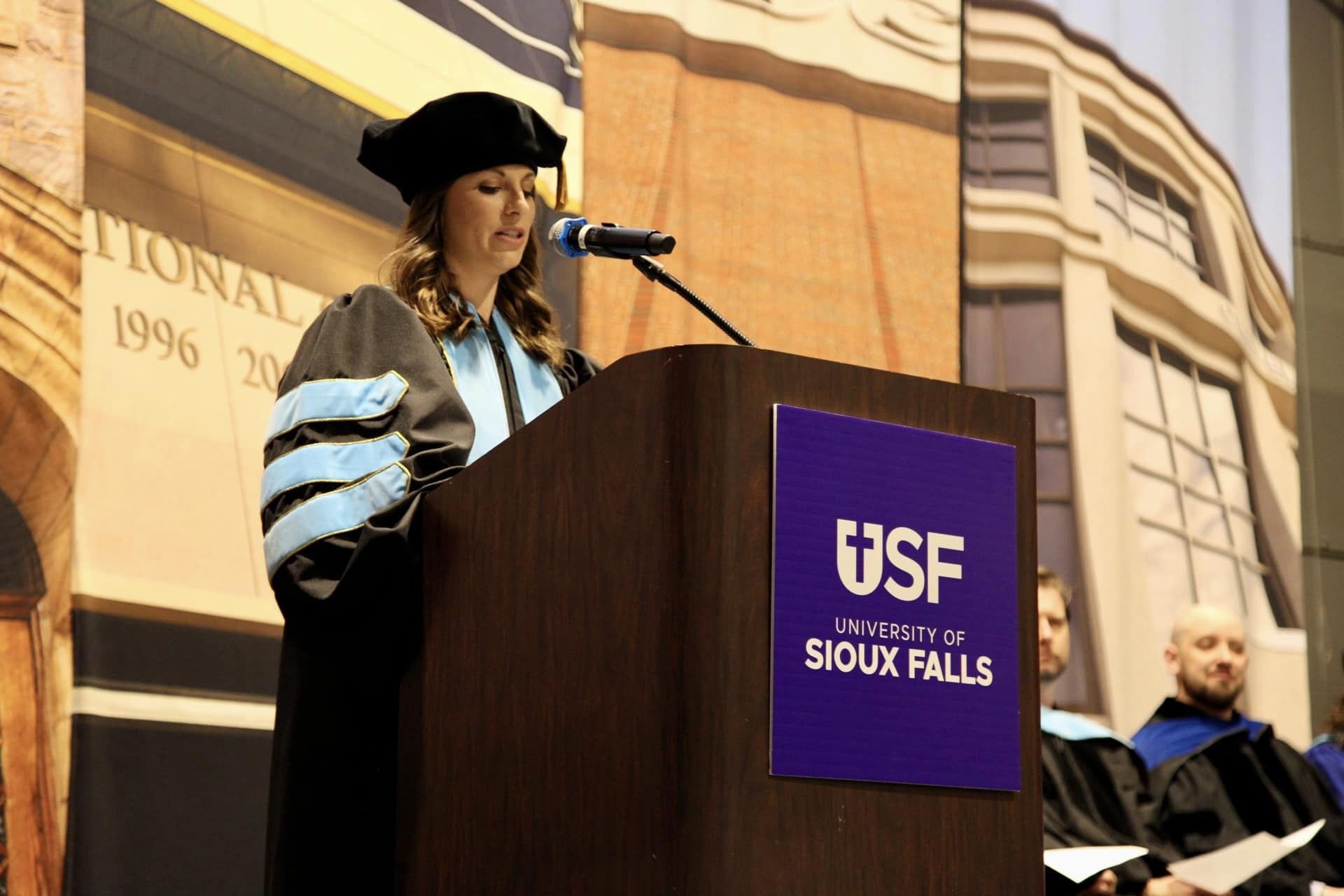 A person wearing academic regalia speaks at a podium with a "University of Sioux Falls" sign. They are addressing an audience as part of an event. In the background, several individuals in similar attire are seated on stage. A building facade is seen behind them.