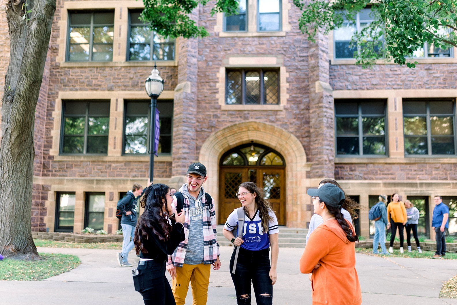 A group of five young adults laugh and converse outside in front of a brick building with large windows. Two are wearing backpacks and one person is holding an iced coffee cup. Other people can be seen in the background near the building.
