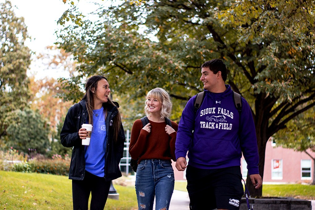 Three college students walk together on a tree-lined campus path. Two women, one holding a coffee cup, and a man wearing a "Sioux Falls Track & Field" sweatshirt are engaged in conversation and smiling. Autumn foliage is visible in the background.