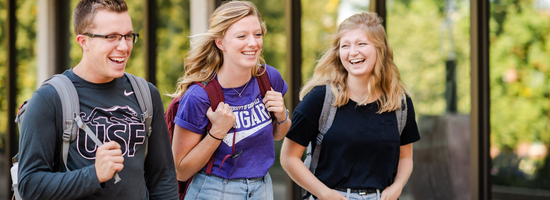 Three individuals walk together outdoors, smiling and carrying backpacks. They appear to be college students. The background shows greenery and a building with large windows. The person in the middle wears a purple t-shirt with "Cougars" written on it.