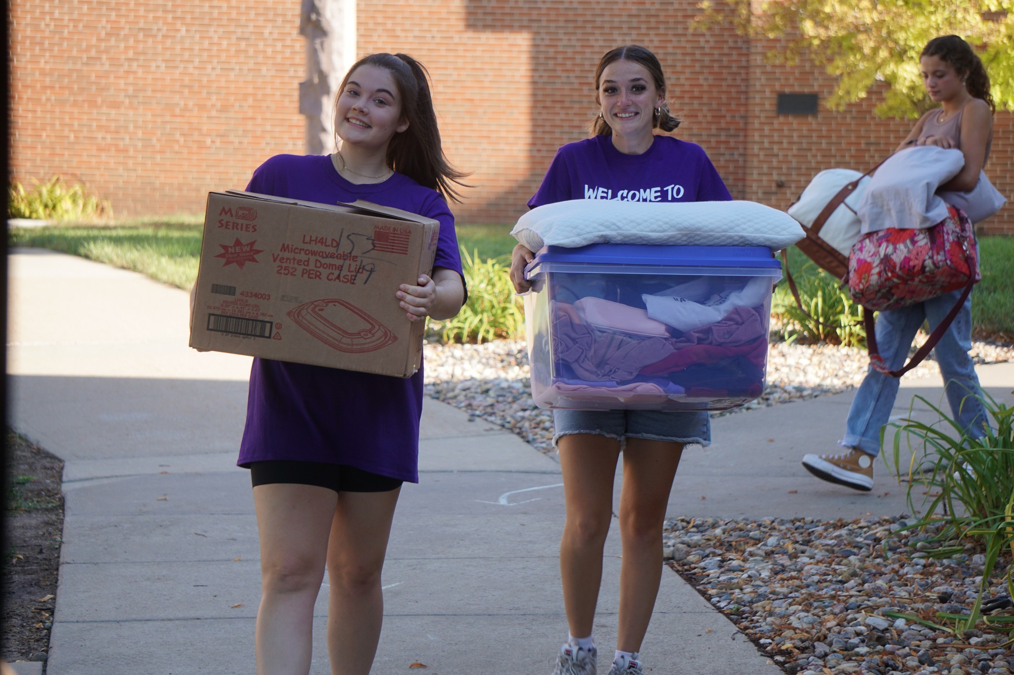 Two smiling young women wearing purple shirts carry belongings, with one holding a cardboard box and the other a large plastic storage bin. They appear to be moving into a dormitory. Another person in the background is carrying bedding.