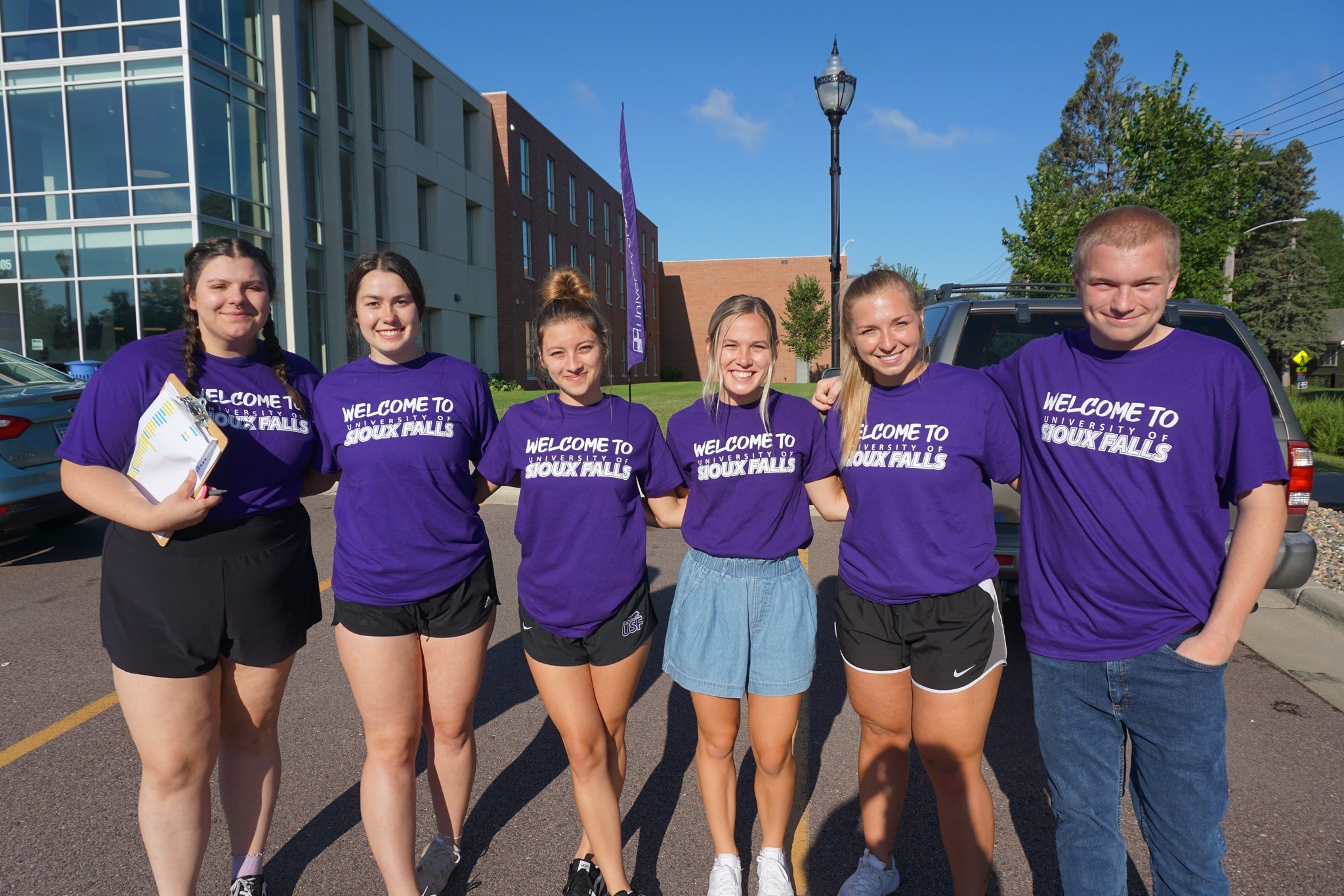 A group of six smiling young adults stands outdoors in bright sunlight, all wearing matching purple T-shirts that read "WELCOME TO SIOUX FALLS." They stand in front of a modern building with large windows, and there are trees and cars in the background.