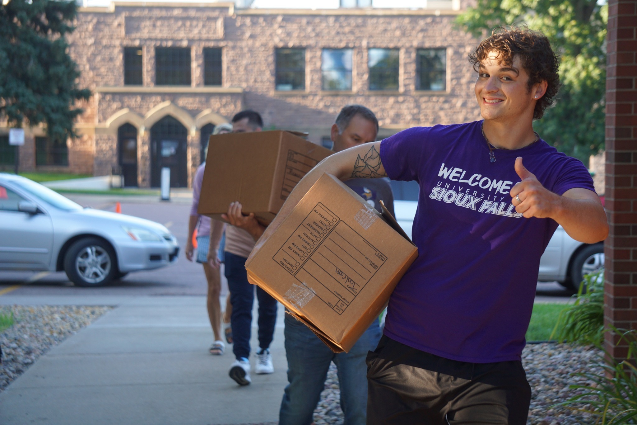 A smiling person wearing a purple "Welcome University of Sioux Falls" shirt gives a thumbs-up while carrying a cardboard box. Several other individuals carrying boxes follow behind. They appear to be moving into a building with a brick exterior.