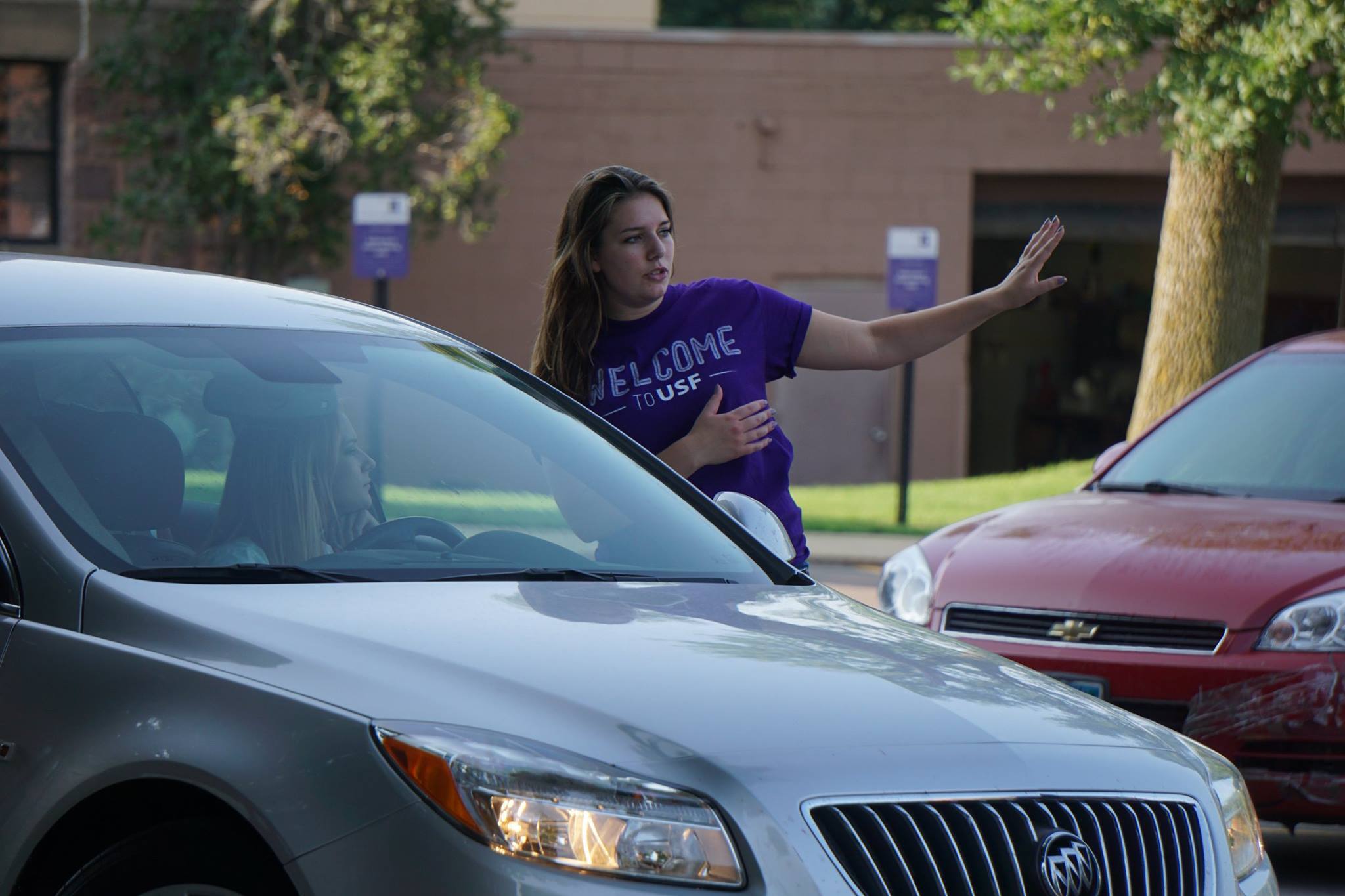 A person wearing a "Welcome to USF" T-shirt is directing a silver car in a parking area, using their right hand to signal the vehicle. The driver is visible through the car window. There are other parked cars and greenery in the background.