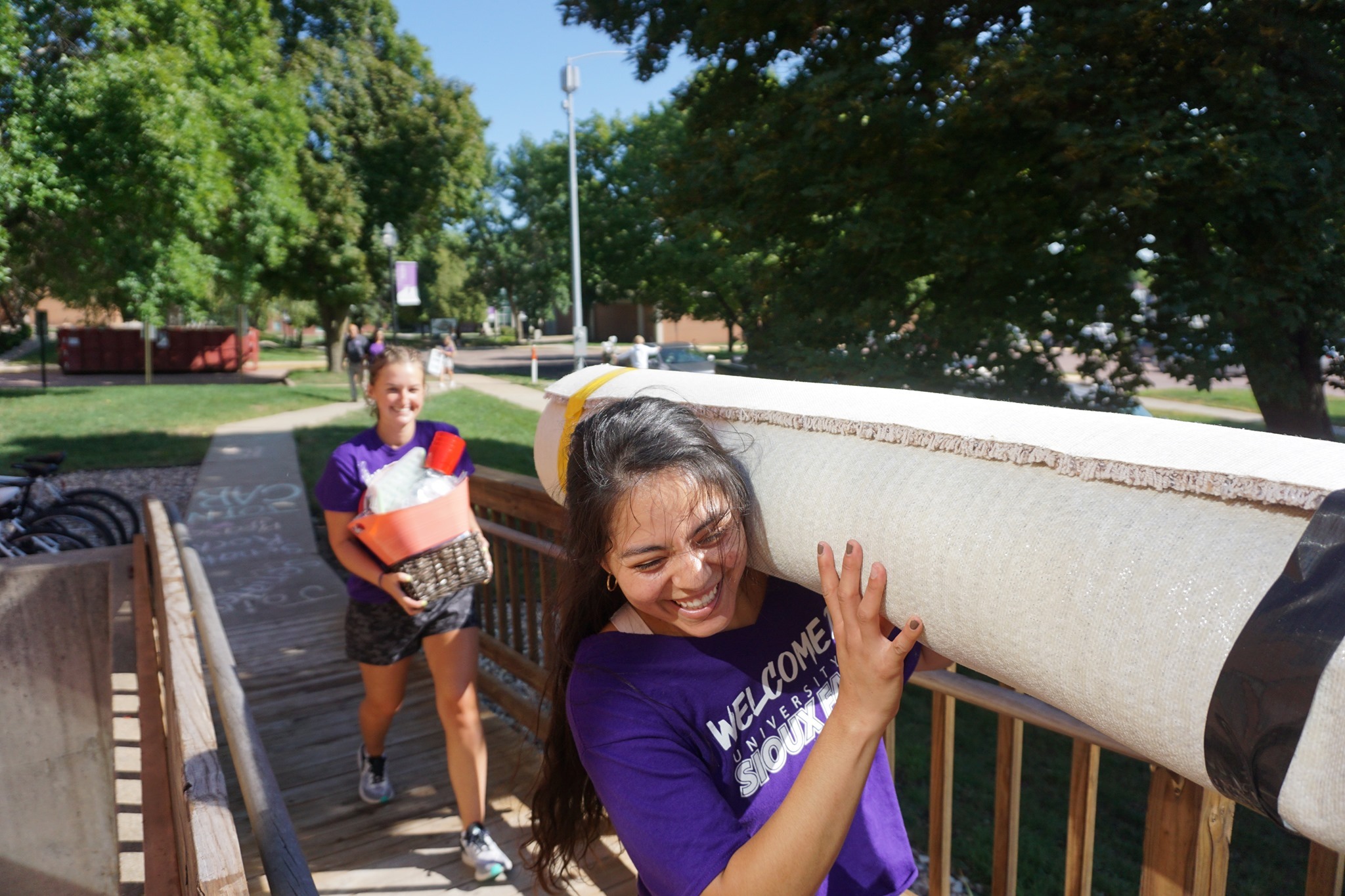Two young people are helping move items outdoors. A person in the foreground is smiling and carrying a rolled-up mattress on their shoulder, while another person in the background is holding a basket filled with various items. Both are wearing purple "Welcome Squad" shirts.