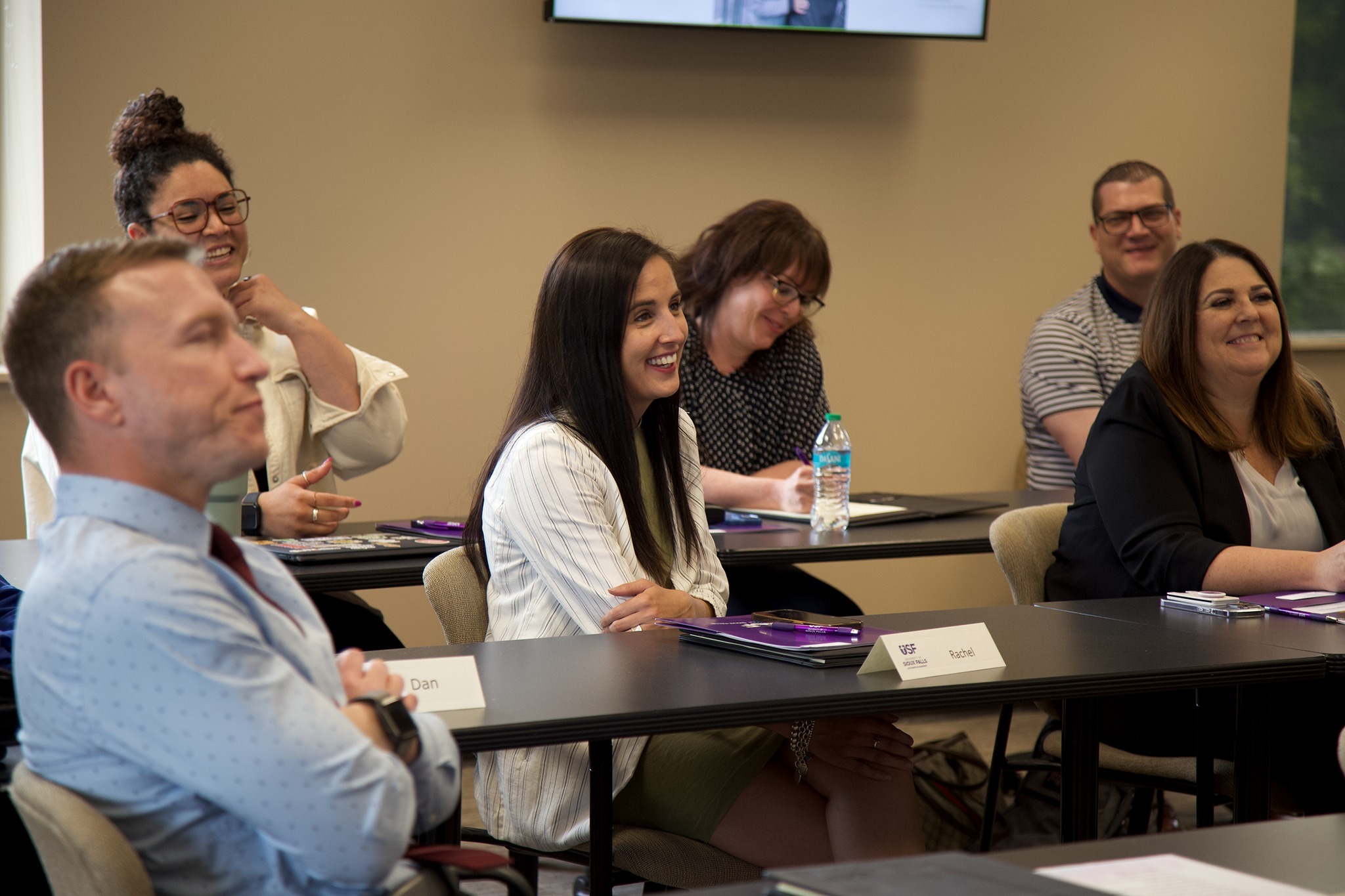 A group of people are seated in a classroom-like setting, attentively looking towards the front of the room. They are smiling and appear engaged. Some of them have nameplates in front of them and notebooks open, suggesting they are attending a lecture or workshop.