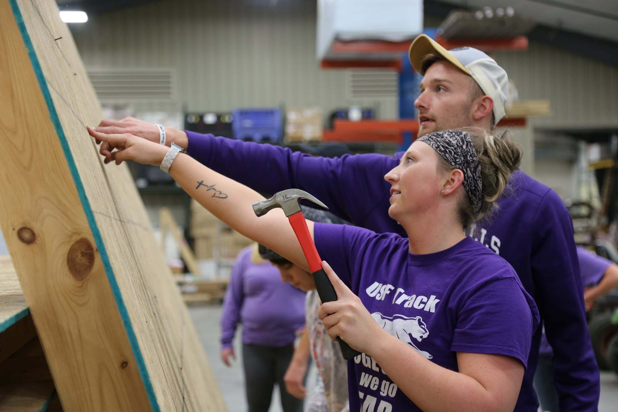 Two individuals are working on a construction project indoors. The person in the foreground, wearing a purple shirt and bandana, holds a hammer and points at a wooden structure. The person behind them, also in a purple shirt and wearing a cap, looks at the same spot.