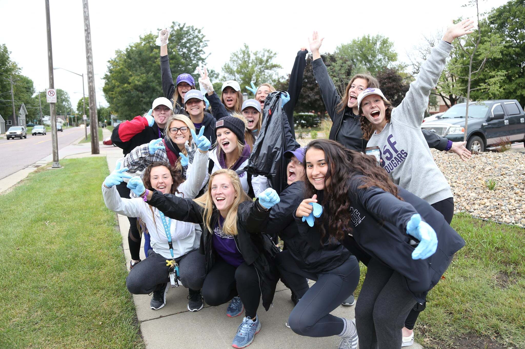 A group of people cheerfully pose for a photo on a sidewalk. They are dressed in casual, weather-appropriate clothing and some wear gloves. In the background, there are trees, a parked car, and residential houses. The group appears to be enjoying the outdoor activity.